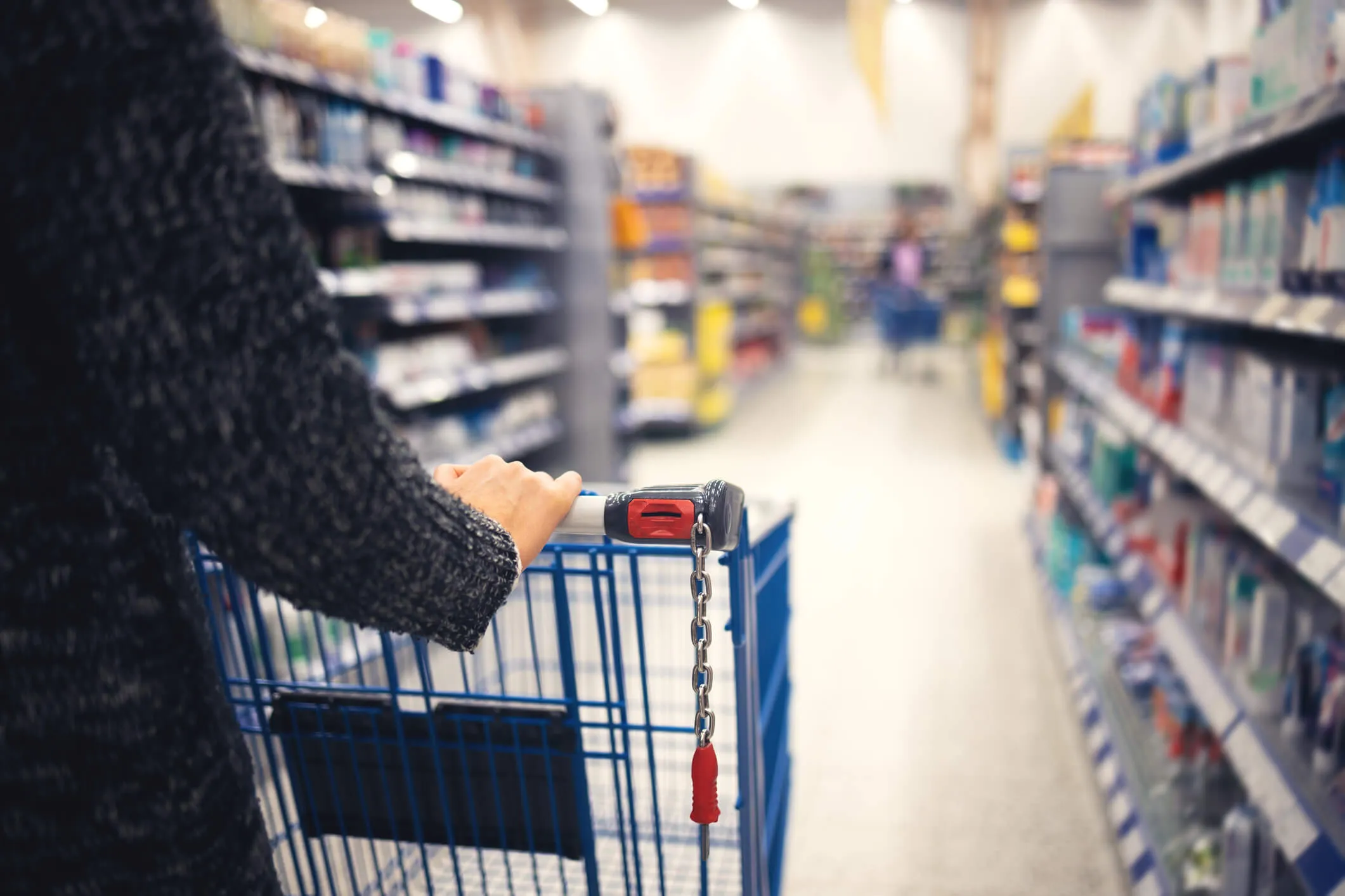 Person pushing a blue shopping cart down a wide grocery store aisle with shelves stocked on both sides.