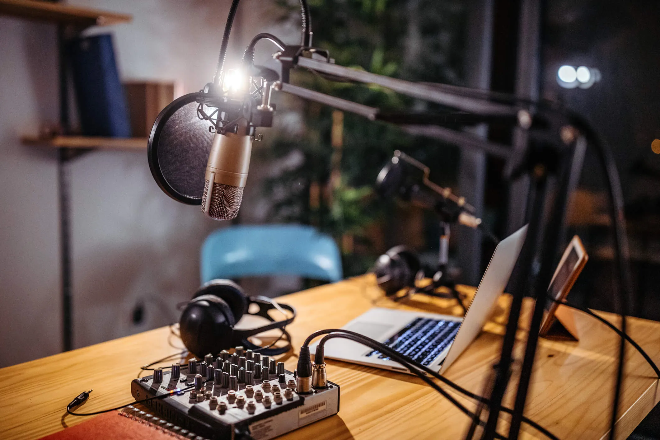 Home podcasting setup with a large condenser microphone on an adjustable arm, audio mixer, headphones, and an open laptop on a wooden desk.