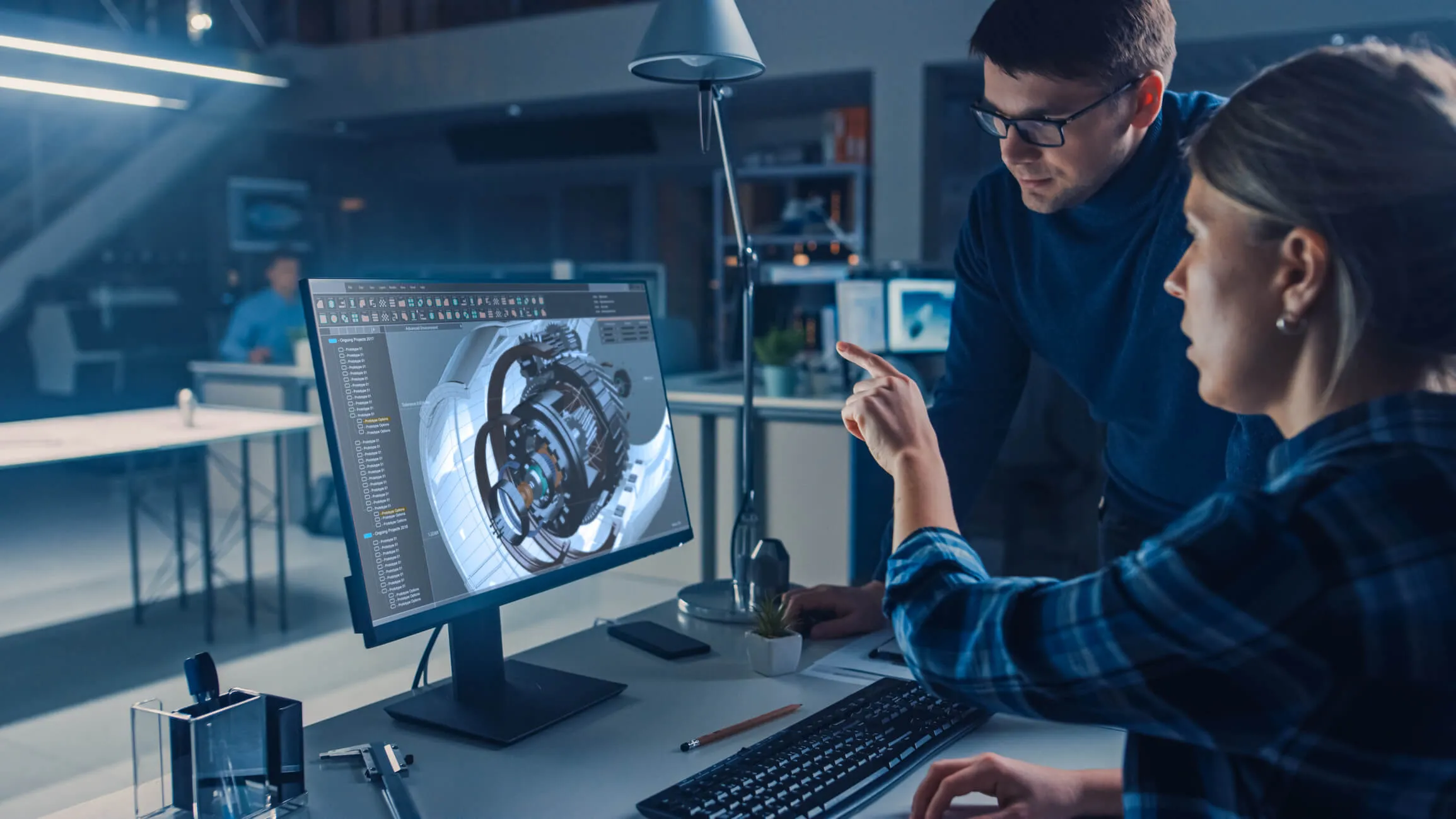 Two engineers in an office collaborate while reviewing a complex 3D mechanical design displayed on a computer monitor.