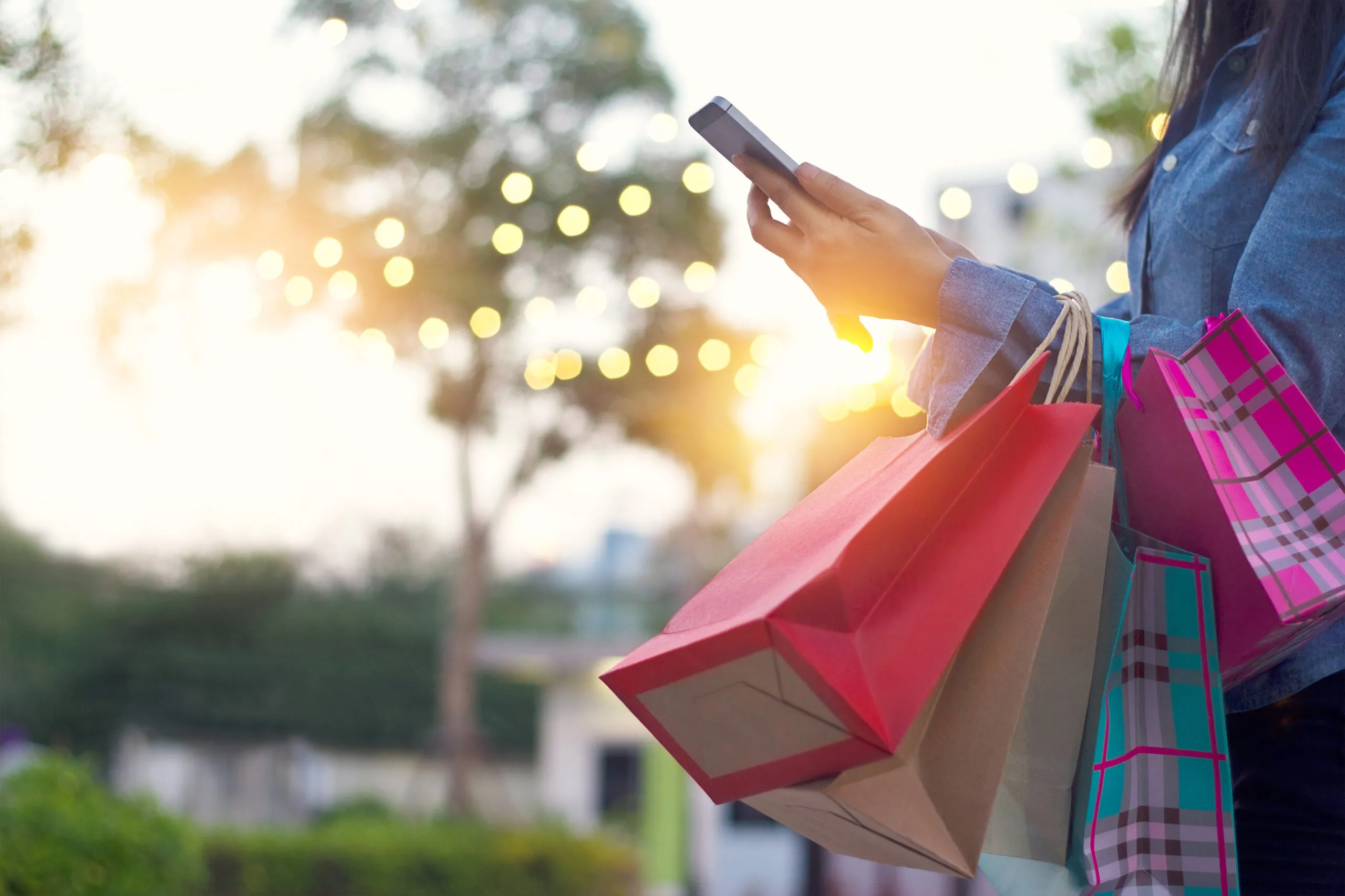 Woman holding multiple colorful shopping bags in one arm and using a smartphone with the other hand outdoors during sunset.