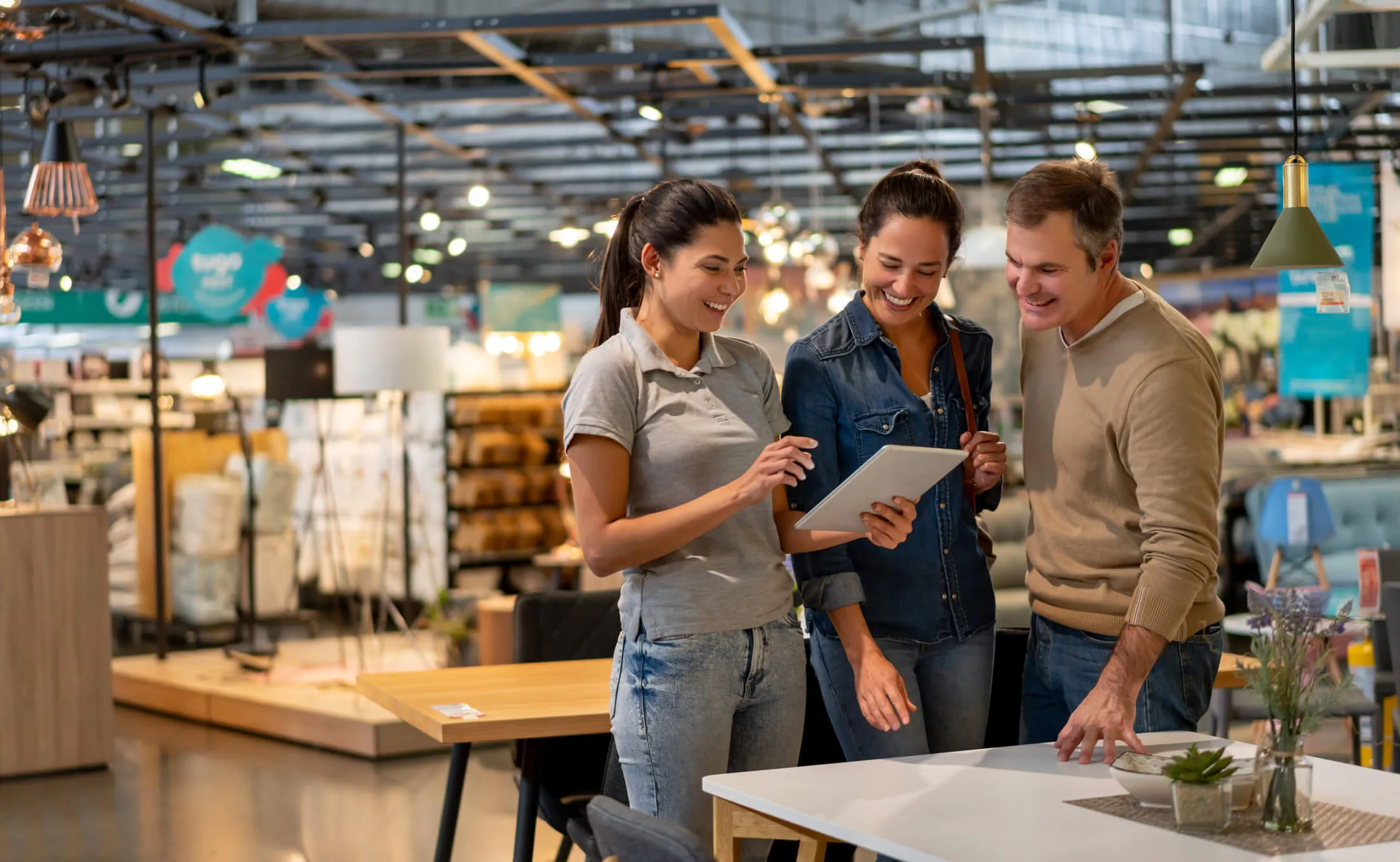 A saleswoman showing a tablet to a smiling couple in a furniture store, with tables, chairs, lamps, and home decor visible in the background.
