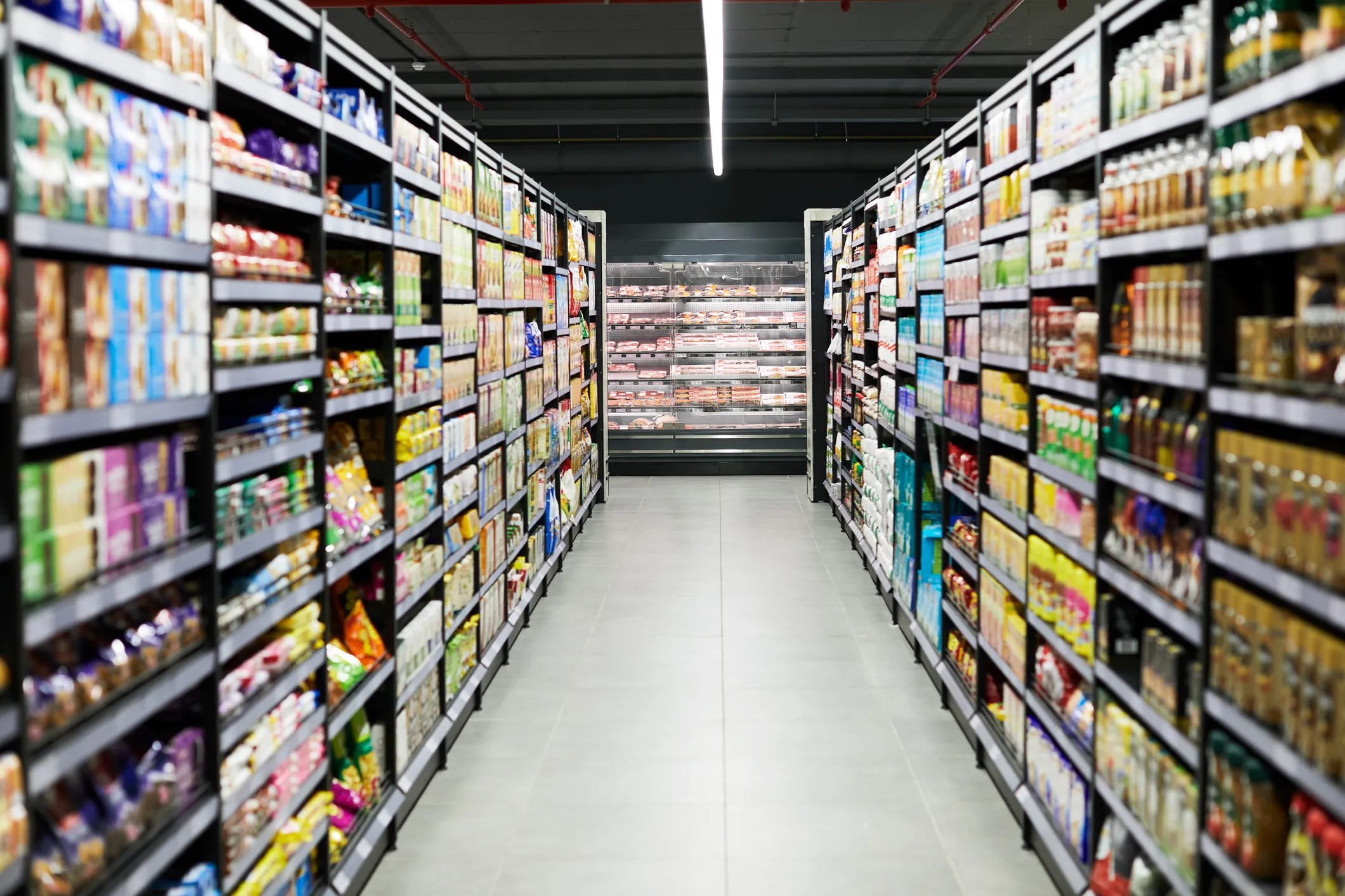 A long grocery store aisle with shelves stocked with various packaged food products, drinks, and household items on both sides, and refrigerated meat products displayed at the end.