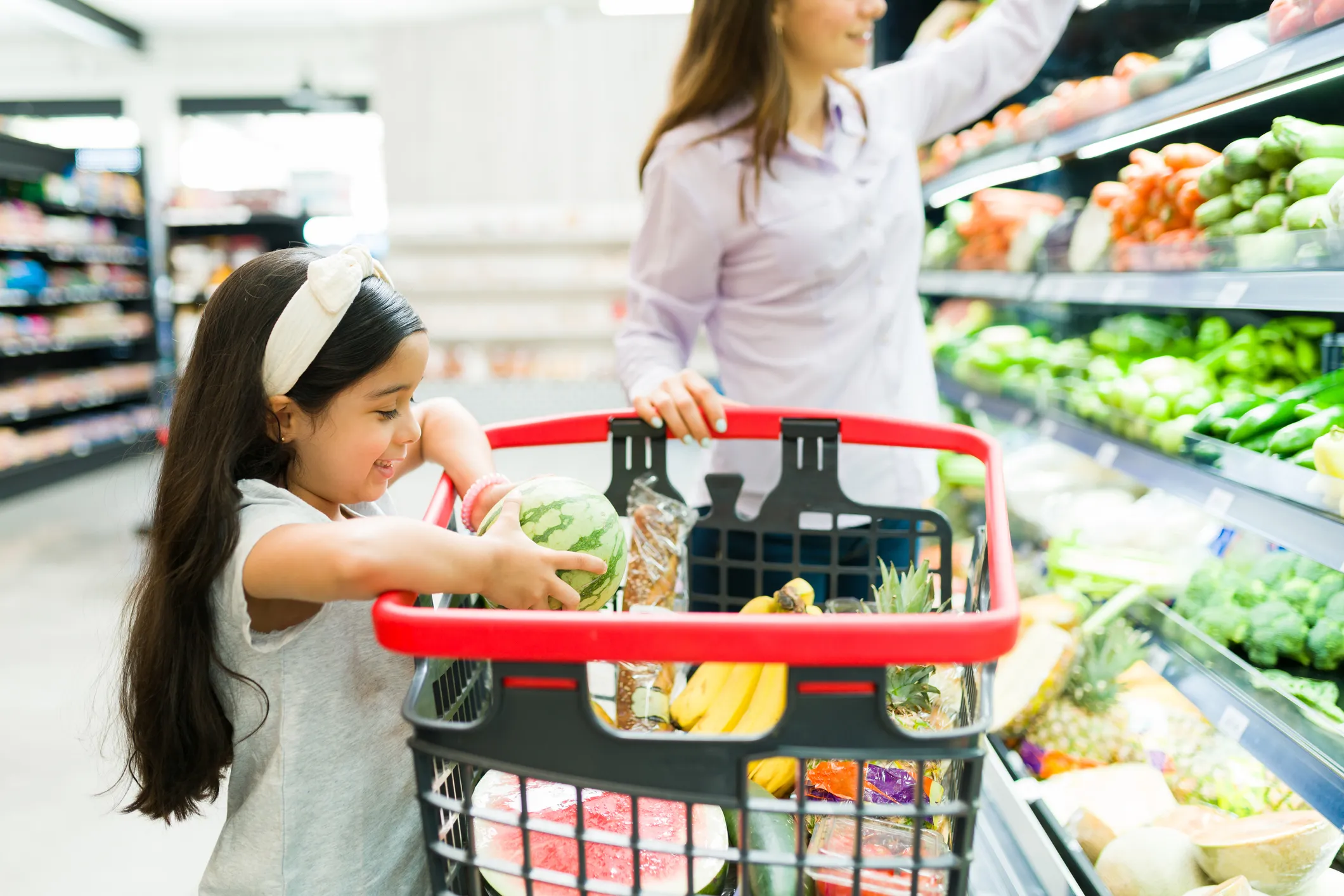 A young girl with a white headband placing a watermelon in a shopping basket filled with fruits and groceries while a woman reaches for produce on a supermarket aisle shelf.