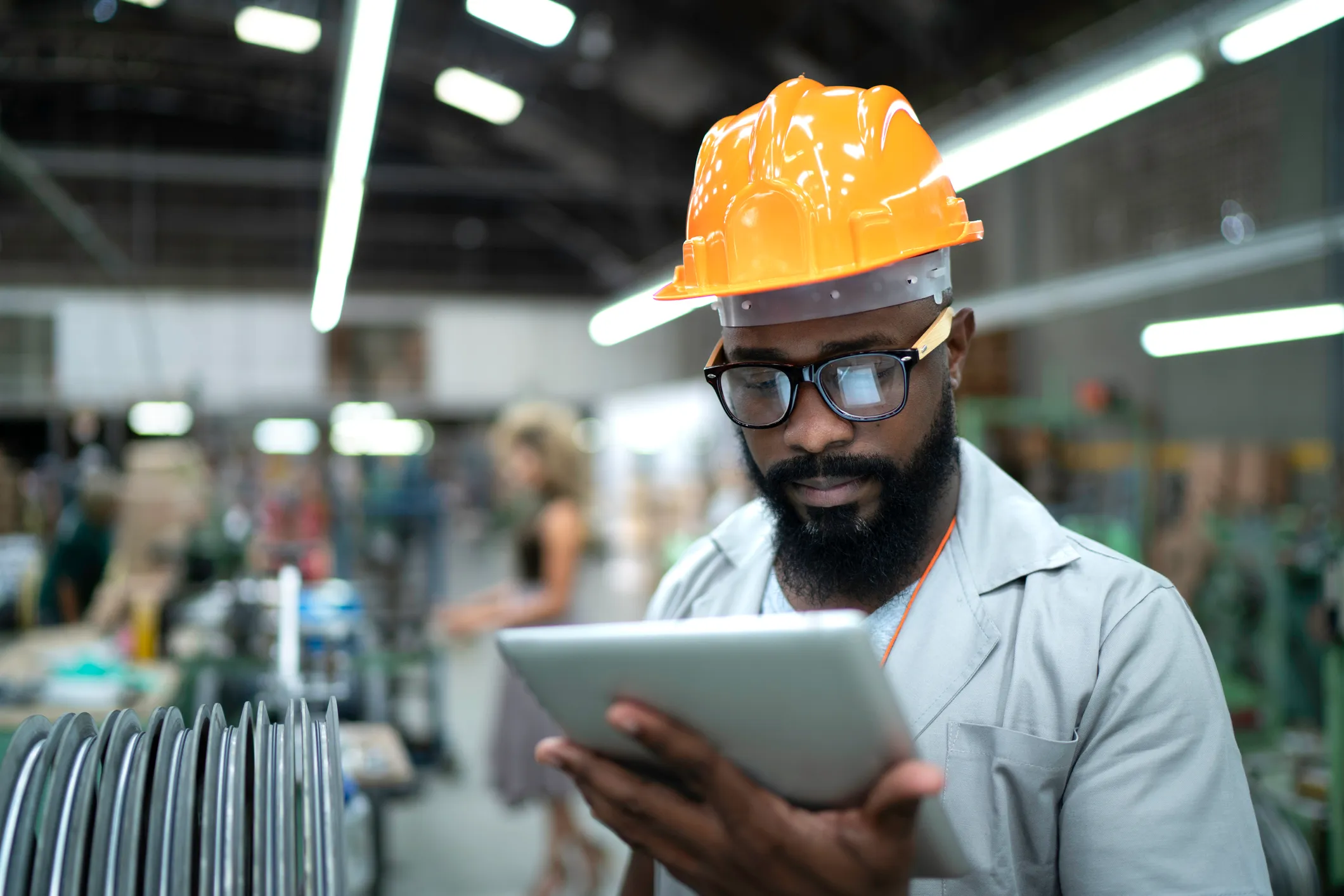 Man with a beard wearing an orange hard hat and glasses, using a tablet in a manufacturing facility.