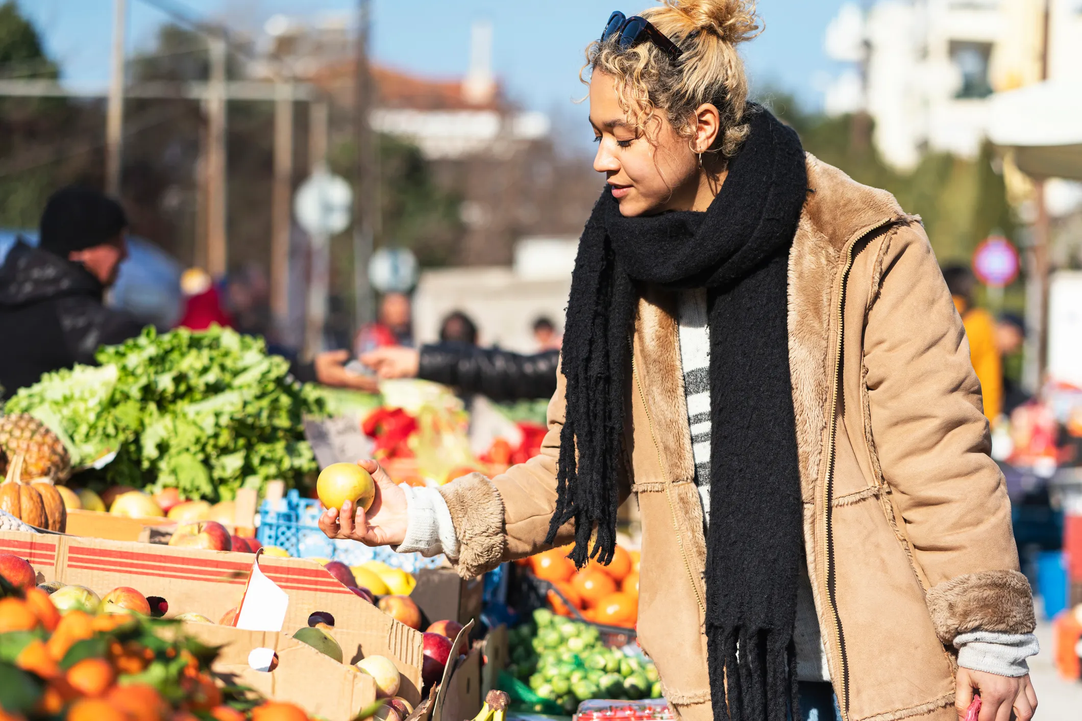 Woman wearing a tan coat and black scarf selecting fruit at an outdoor market stall filled with fresh produce including apples, lettuce, oranges, and pineapples.