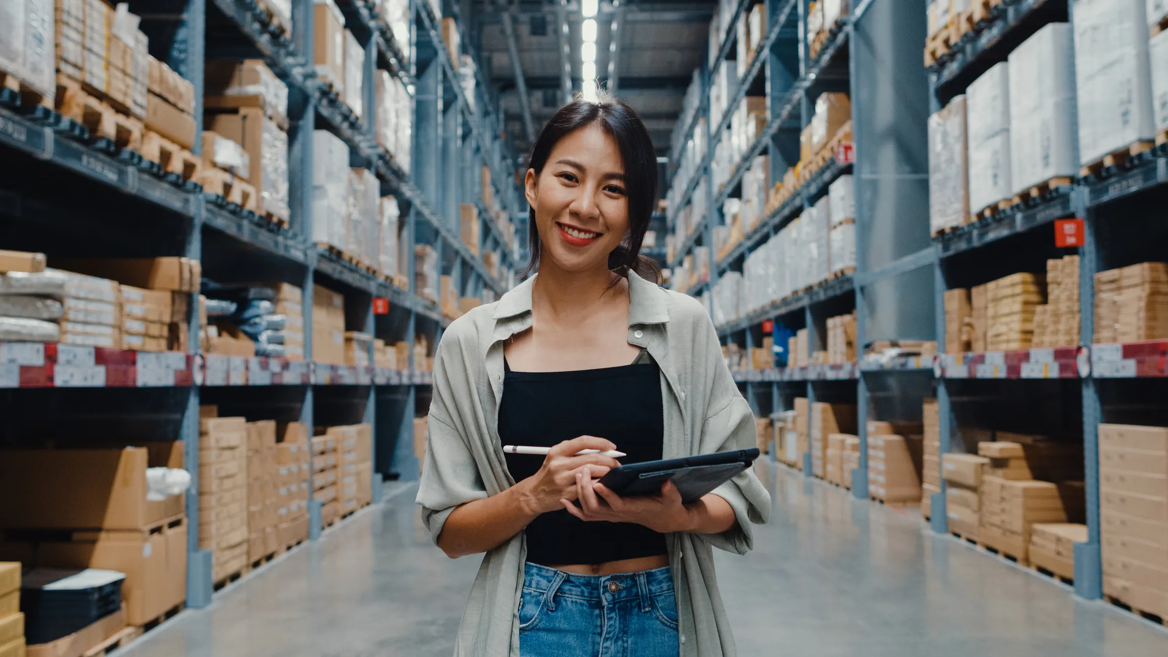 Smiling woman standing in a warehouse aisle holding a tablet and stylus, with high shelves stocked with boxes on both sides.