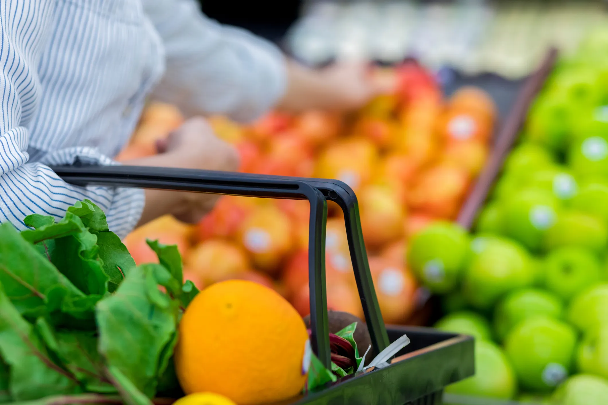 Close-up of a shopping basket filled with fresh leafy greens and an orange, held by a person wearing a striped shirt in a produce section with apples and other fruits in the background.