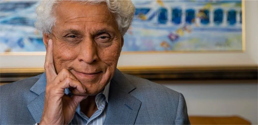 Portrait of an older man with white hair and warm skin tone, smiling gently with his hand touching his face, wearing a blue blazer and striped shirt with a colorful painting in the blurred background.