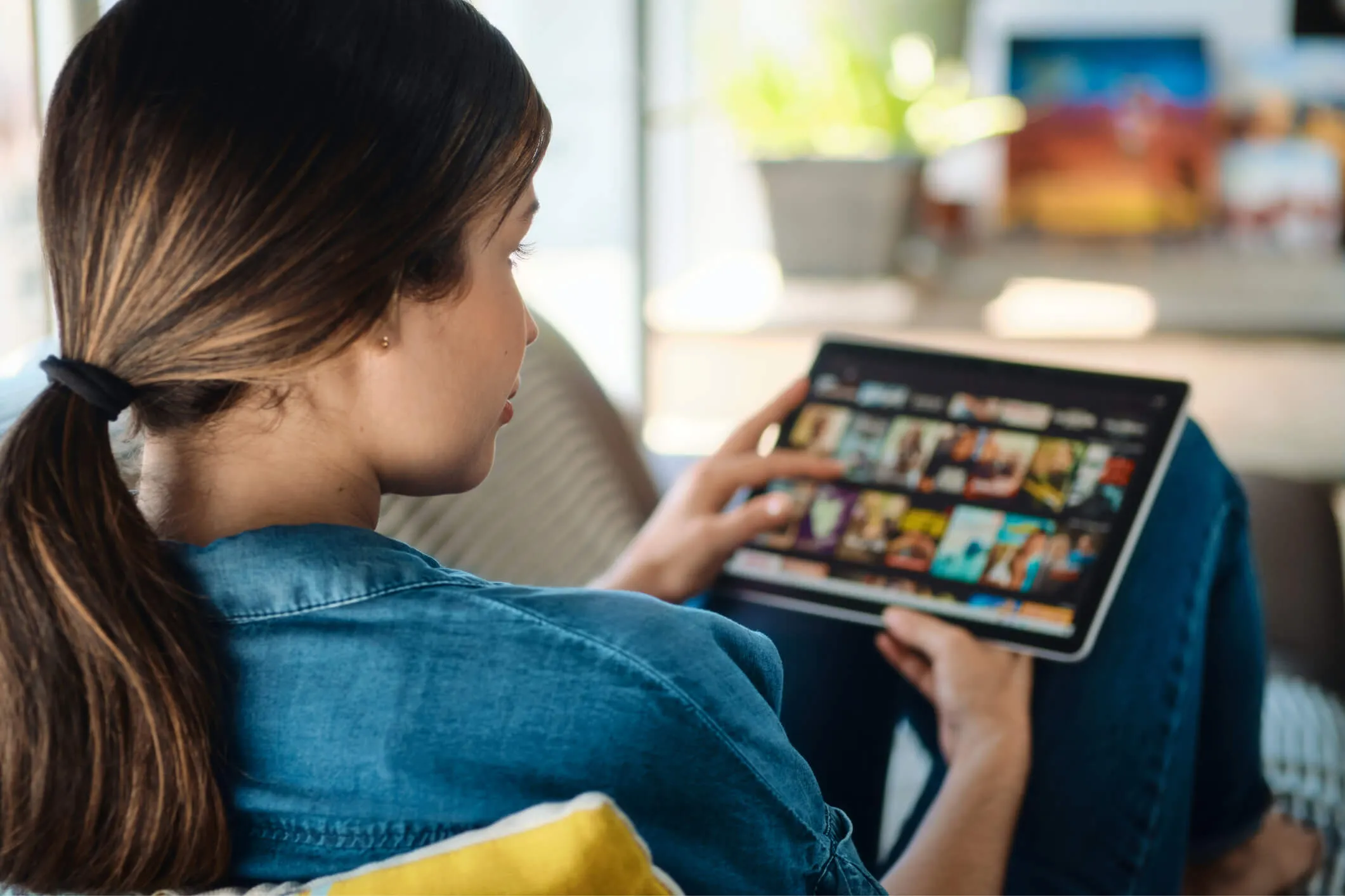 A woman with a ponytail and wearing a blue shirt is sitting and browsing a tablet displaying a streaming service interface with various show thumbnails.