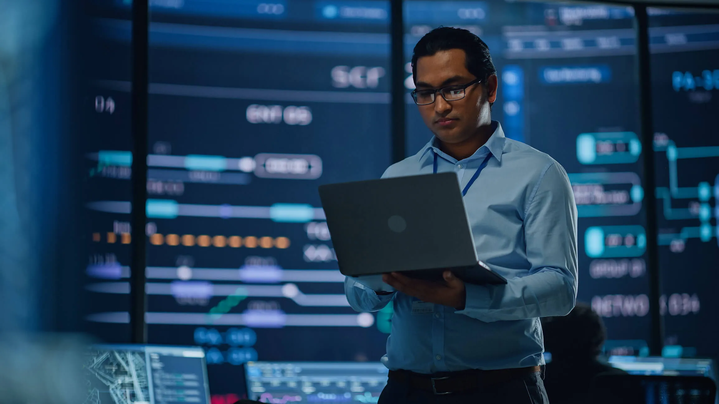 Man wearing glasses and a blue shirt holding a laptop, working in a high-tech control room with large digital display screens showing complex data and graphics.