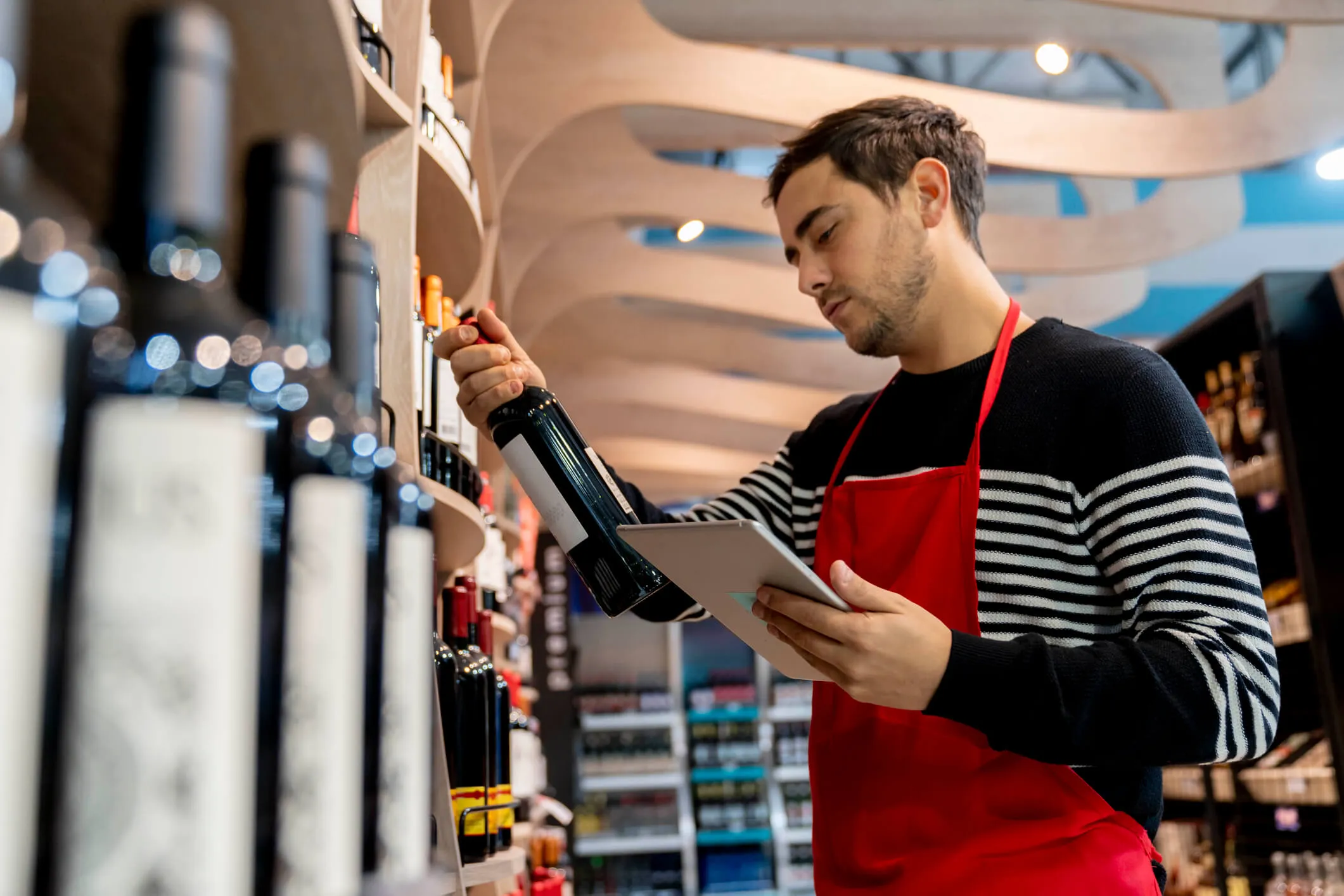 A man wearing a red apron and black striped sweater examines a wine bottle in a store while holding a tablet, standing in front of wooden shelves stocked with wine bottles.