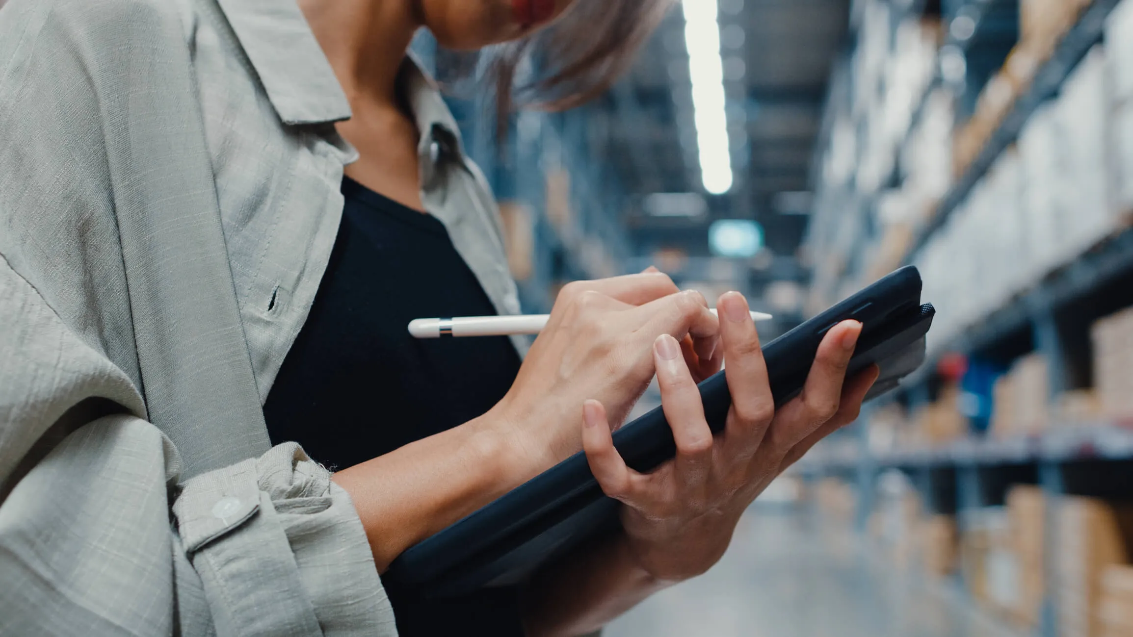 Person in a warehouse holding a tablet and stylus, using the tablet with shelves of boxes blurred in the background.