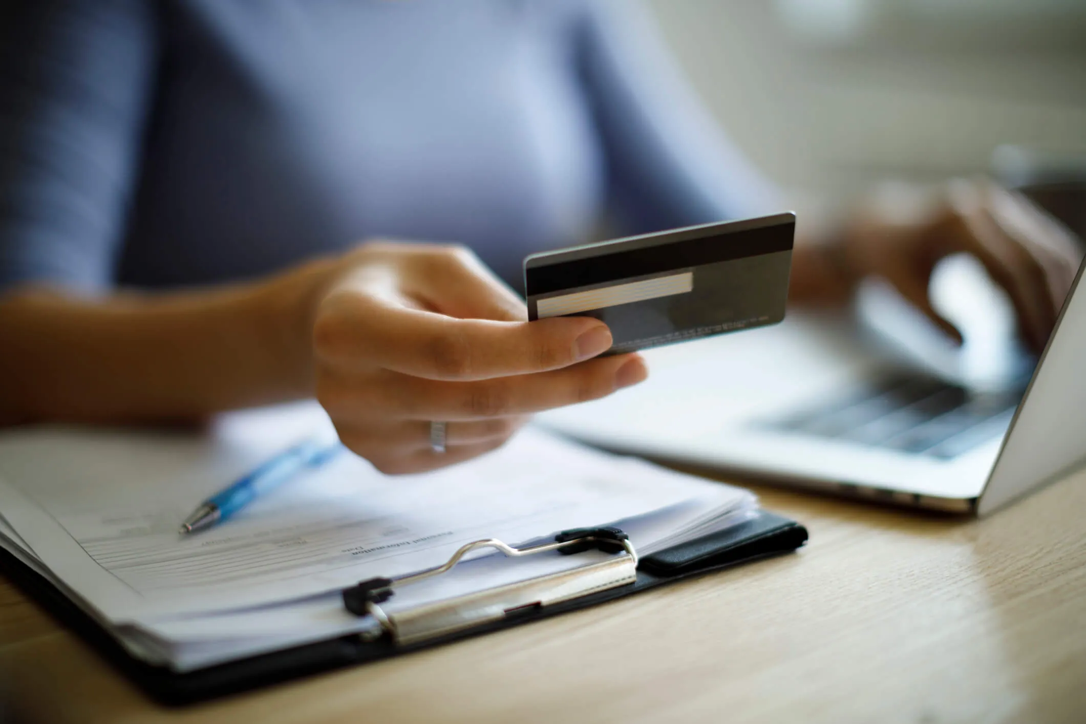 Person holding a credit card in one hand and typing on a laptop with the other, next to a clipboard with papers and a pen on a wooden table.