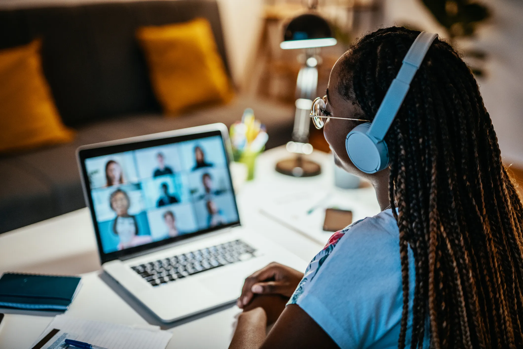 Person with braided hair wearing headphones and glasses participates in a video conference on a laptop, sitting at a desk with notebook, pen, and lamp in a cozy room.