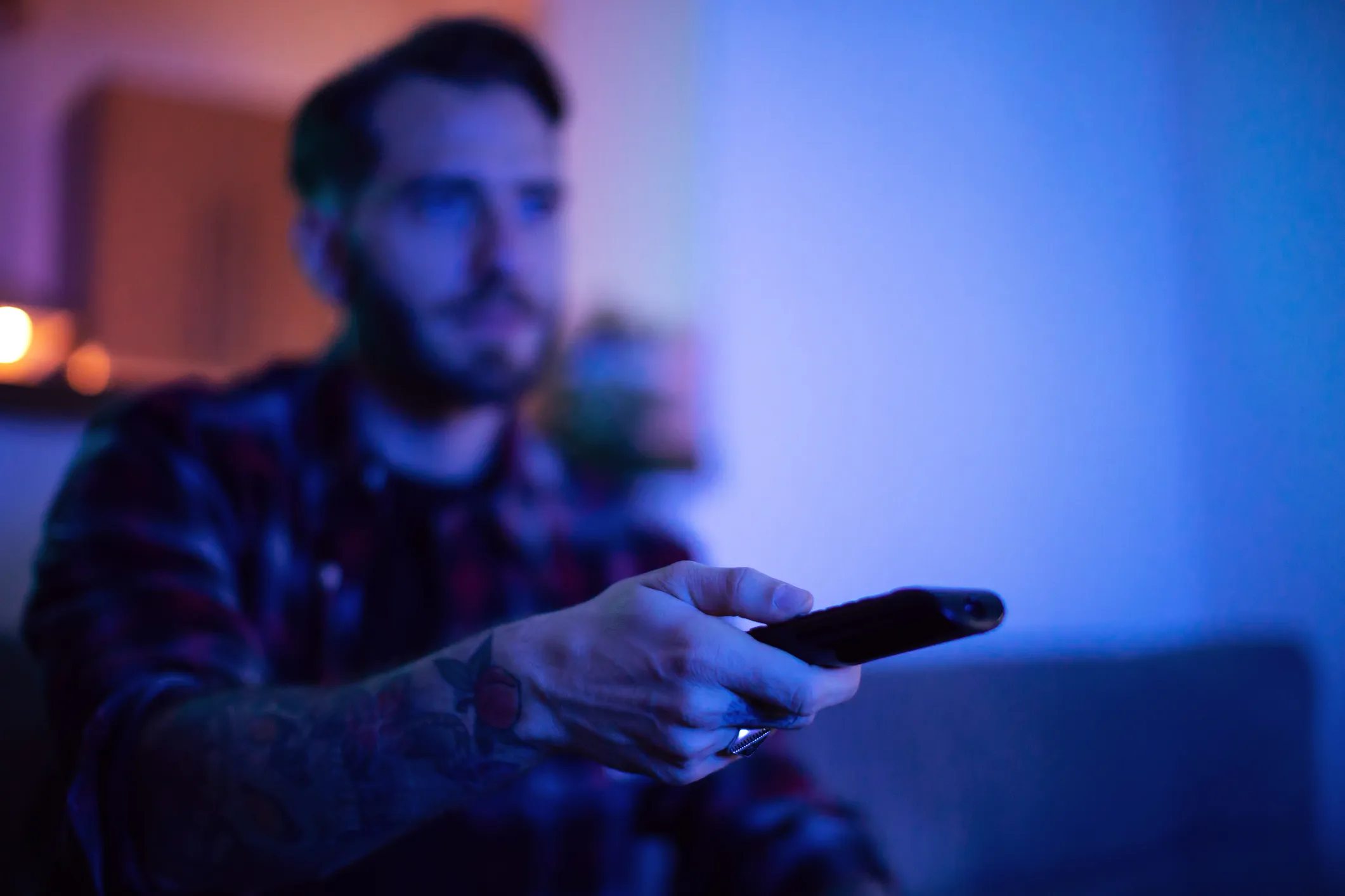 Man with a tattooed arm holding a remote control, illuminated by blue light in a dim room.