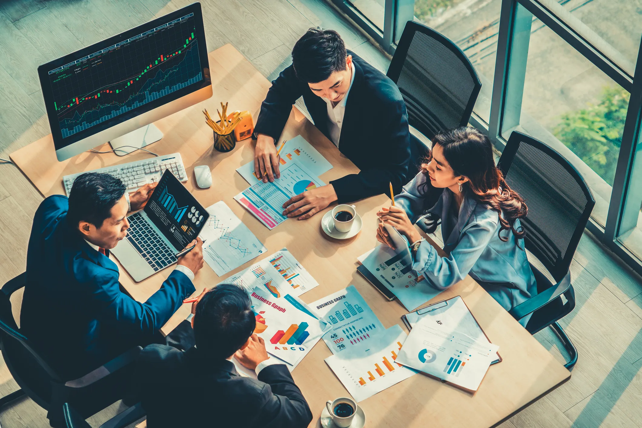 Four business professionals in a meeting around a table with charts, graphs, and laptops showing financial data, discussing strategies in a bright office with large windows.