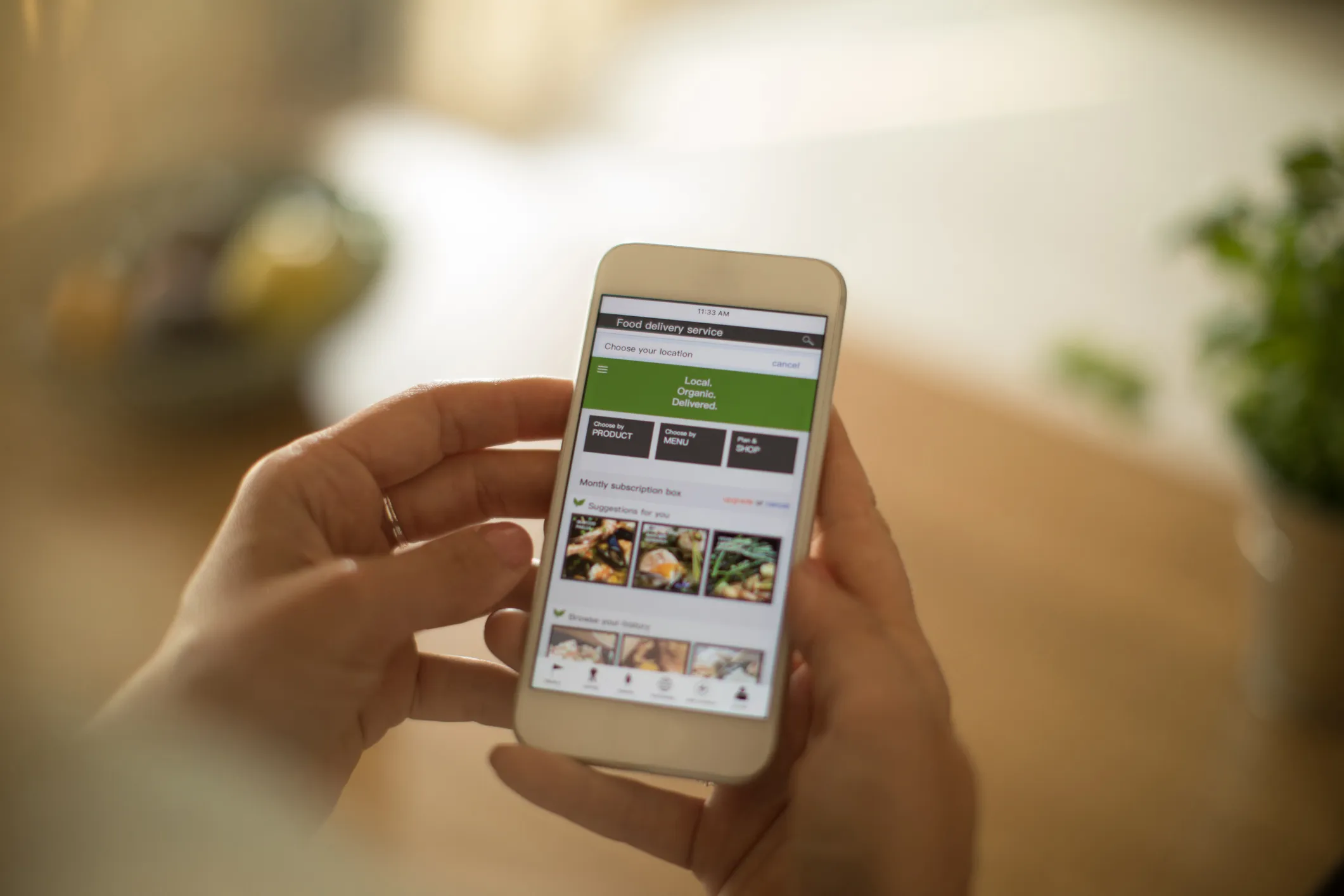 Person holding a smartphone displaying a food delivery service app with options to choose by product, menu, or shop, and sections for monthly subscription box suggestions and browsing order history.