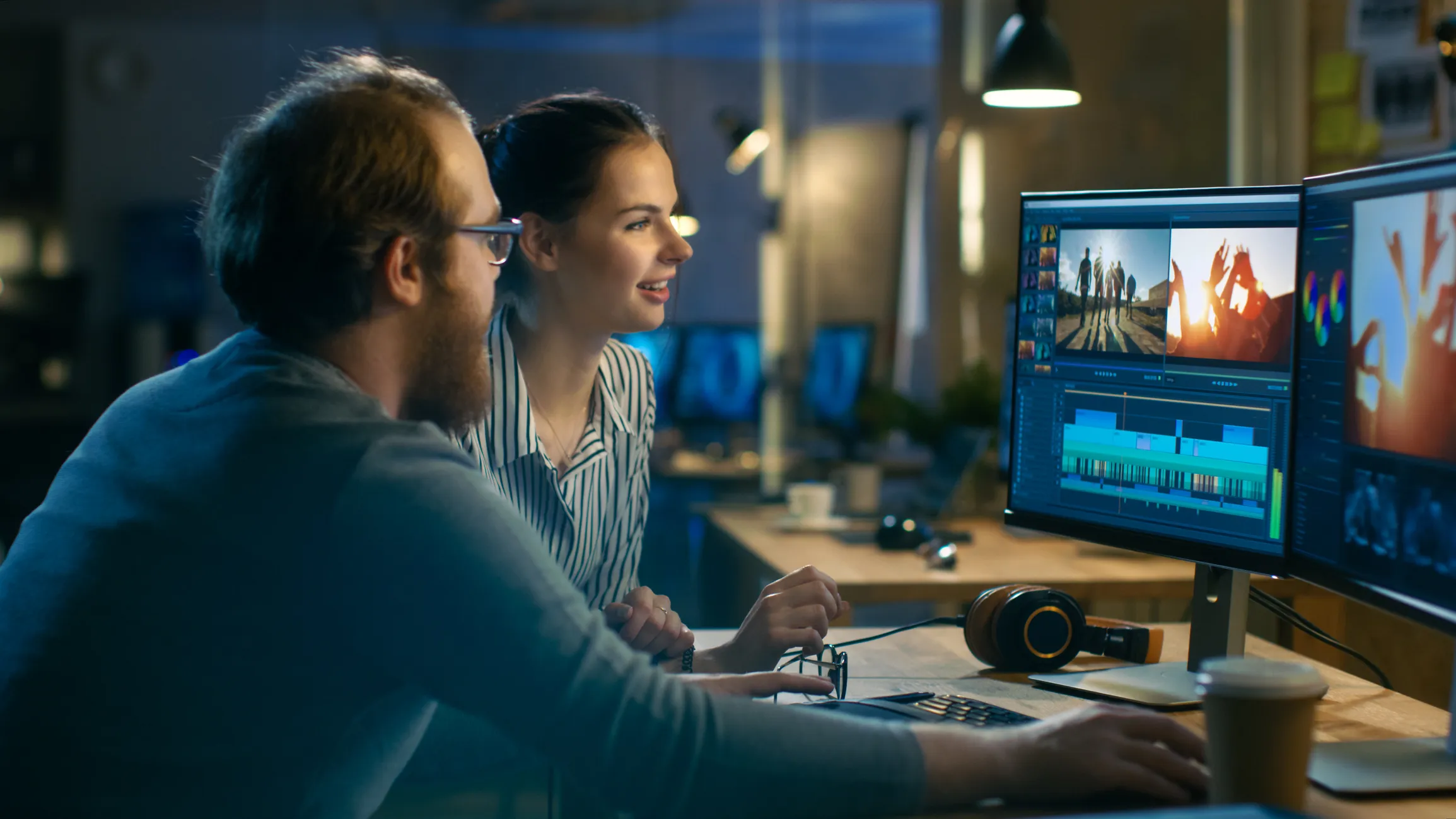 Two people, a man and a woman, working together on video editing on a dual monitor setup in a dimly lit room, with headphones and coffee cup on the desk.