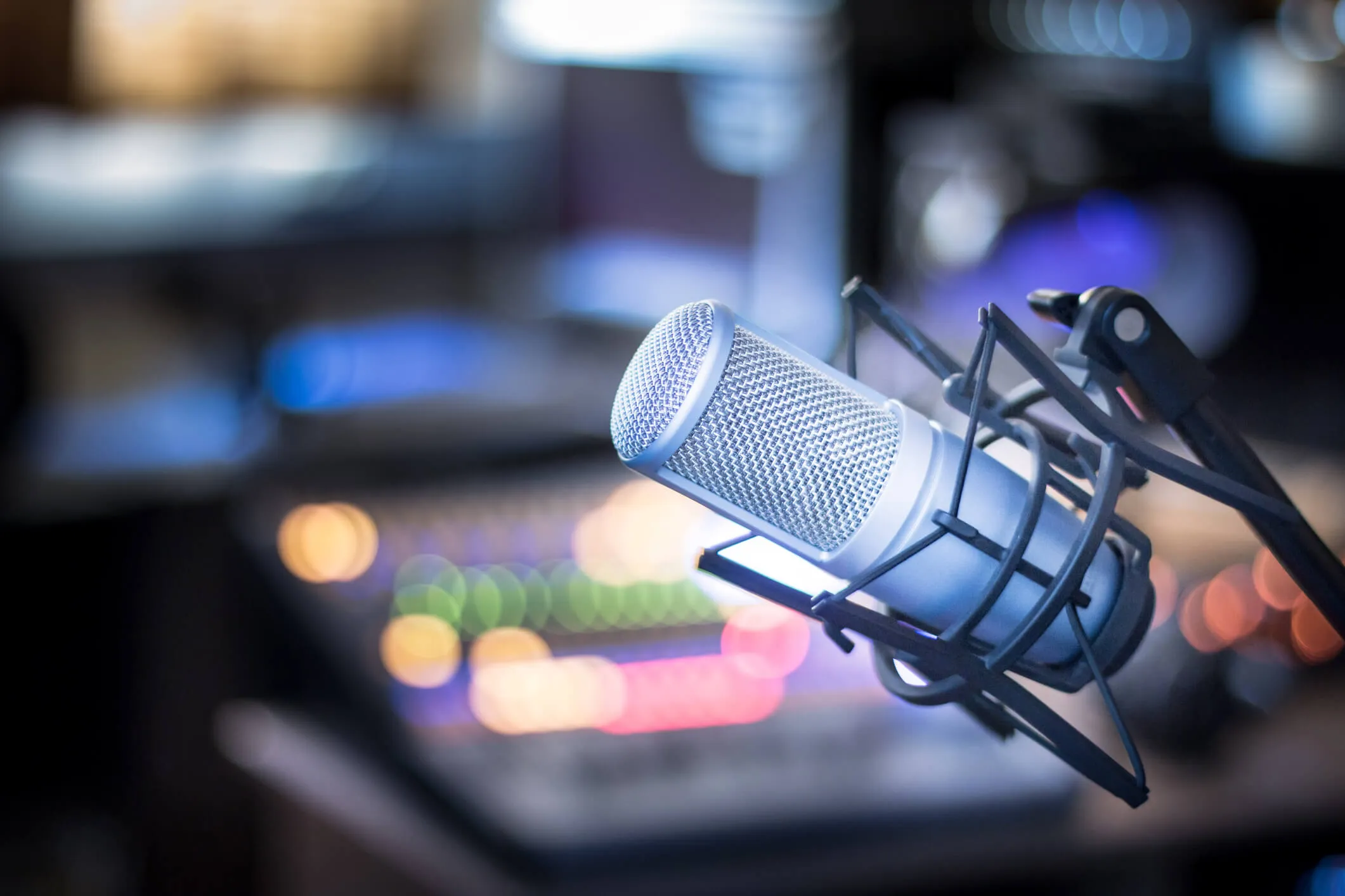 Close-up of a silver condenser microphone with a shock mount in front of a blurred mixing console with colorful lights in a recording studio.