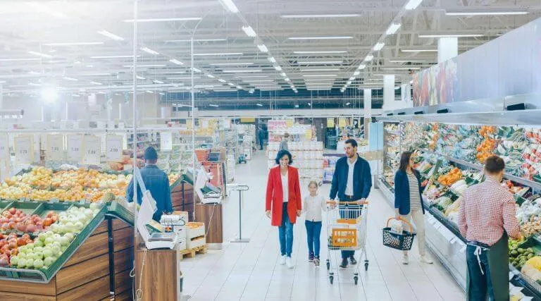 A family of three, including a man pushing a shopping cart, a woman, and a child, walk through a bright supermarket aisle with fresh fruits and vegetables on display, while other shoppers and store workers are nearby.