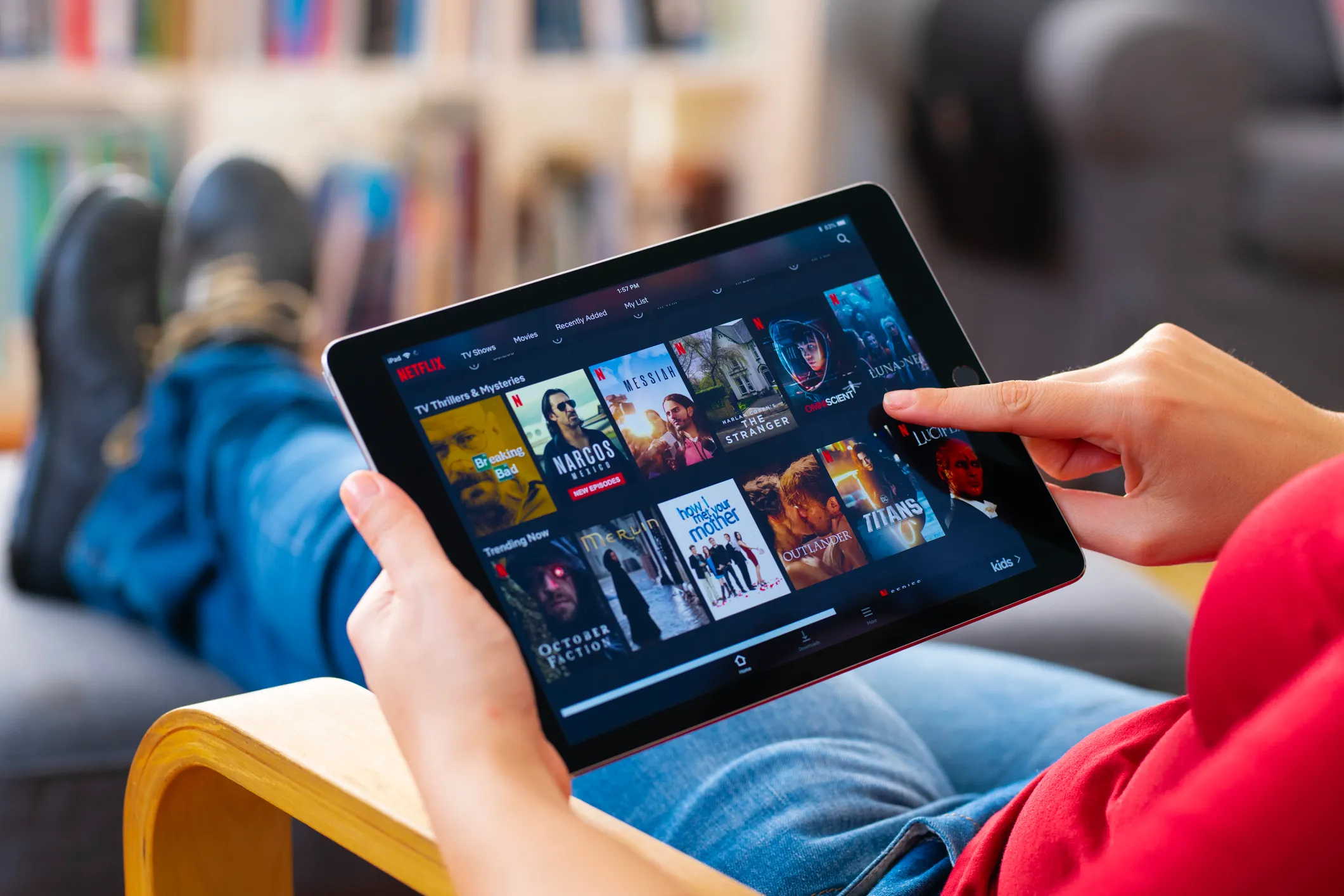 Person browsing Netflix TV shows and movies on a tablet while sitting on a chair with legs stretched out, finger pointing at the screen.