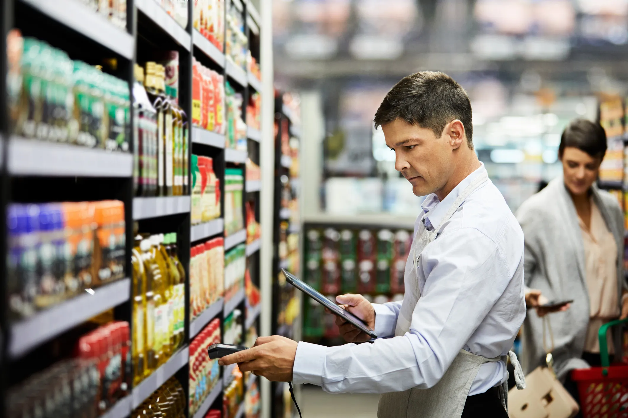 Man wearing a light blue shirt and apron scanning products on a grocery store shelf using a handheld barcode scanner and a tablet, with a woman holding a phone and shopping basket in the background.