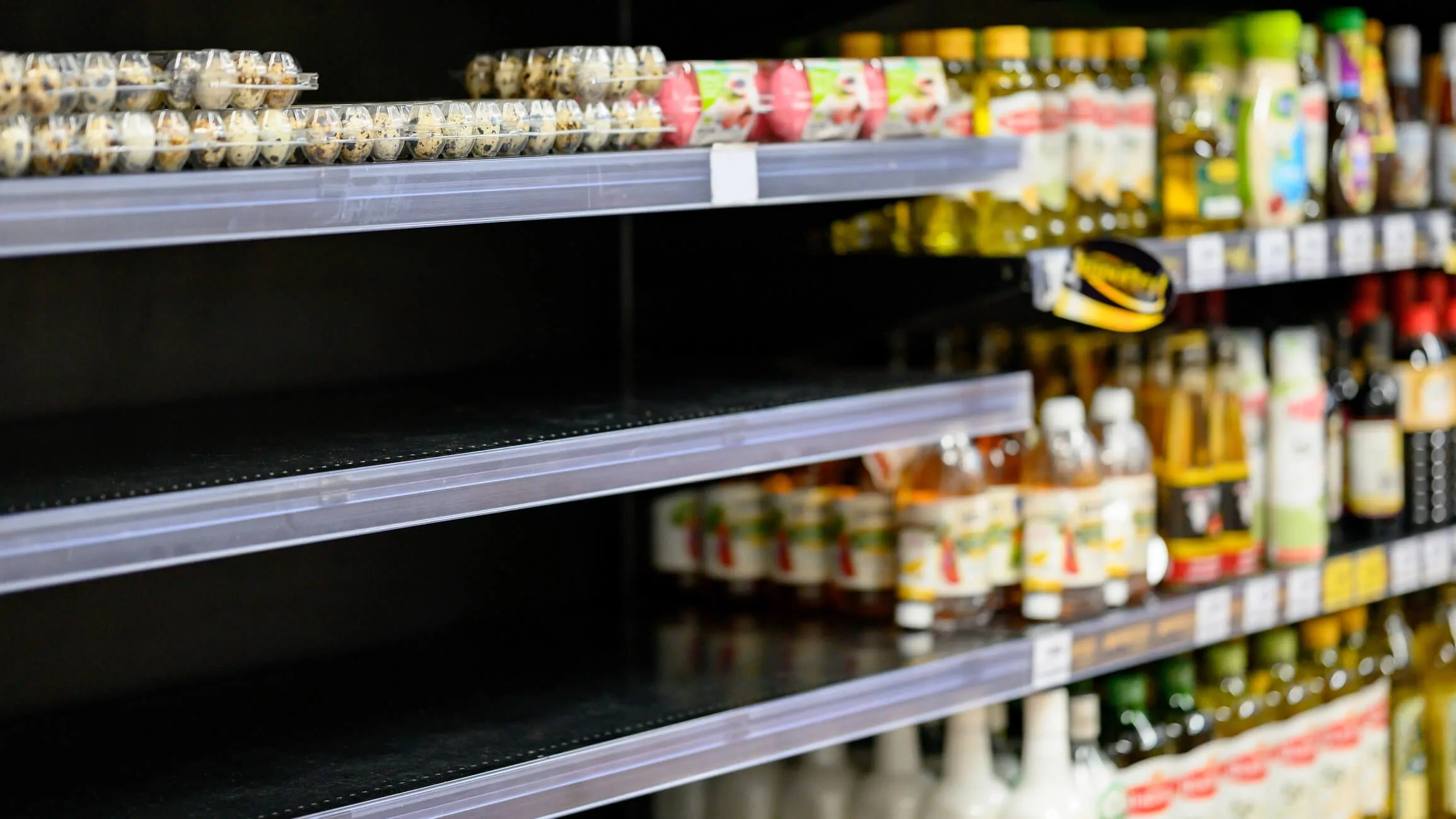 Empty shelves in a grocery store with a few remaining products, including eggs in clear cartons on the top shelf and various bottled items on lower shelves.