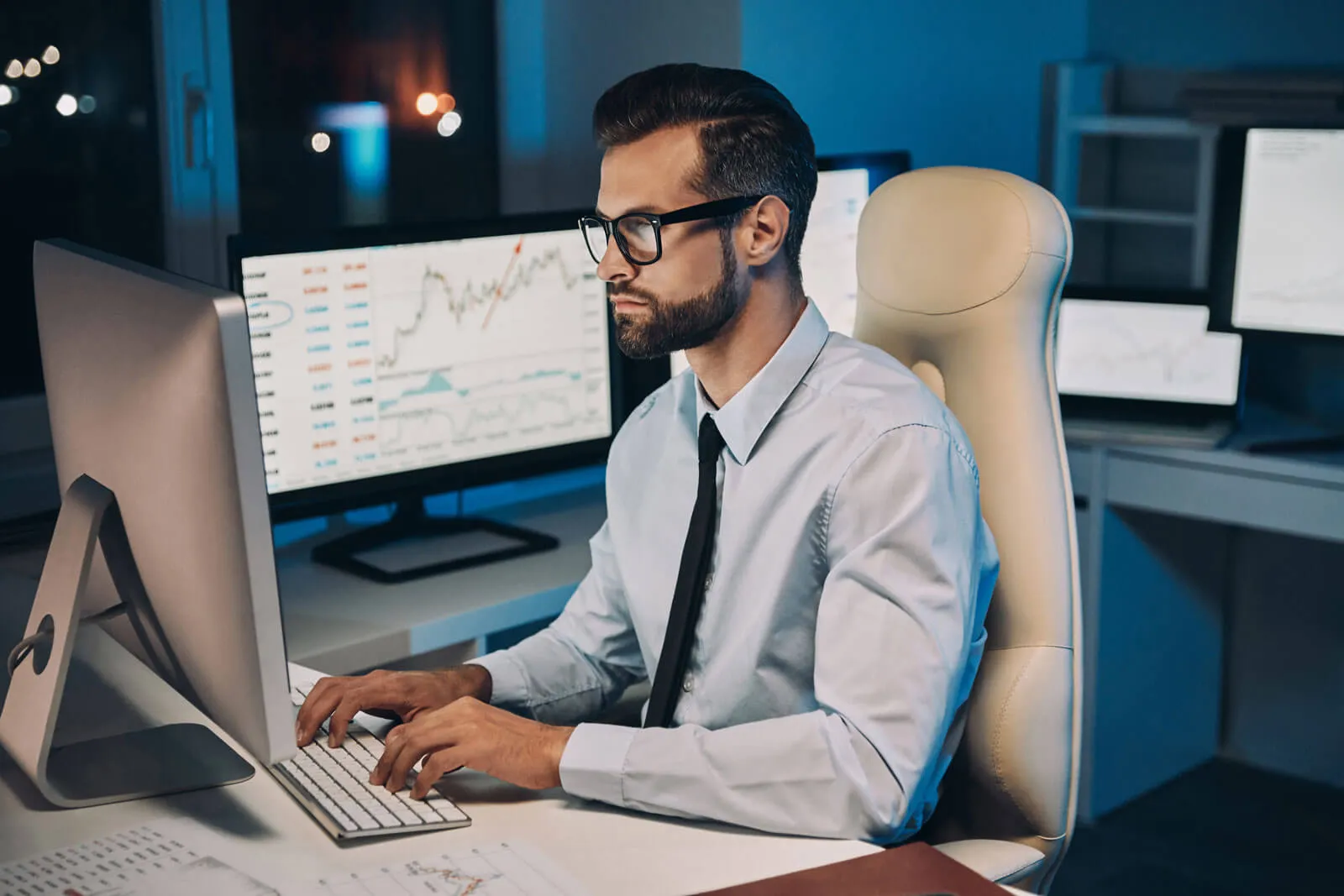 Man in glasses and a dress shirt with a black tie working on a desktop computer in an office with multiple monitors displaying financial charts and data.