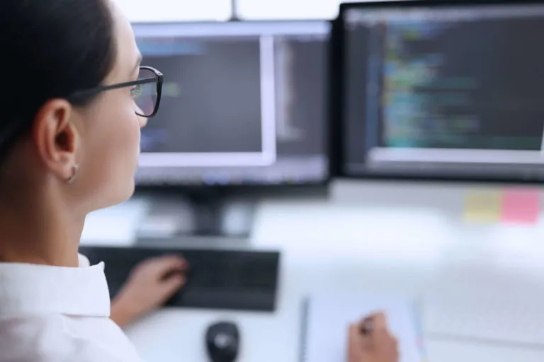 Person wearing glasses working on a computer with two monitors displaying code, taking notes in a notebook.
