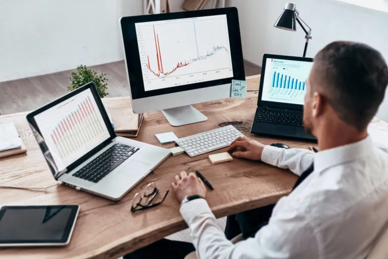 Man in white shirt working at a wooden desk with three screens displaying various data charts and graphs, including line and bar charts, surrounded by office items like glasses, a tablet, and sticky notes.