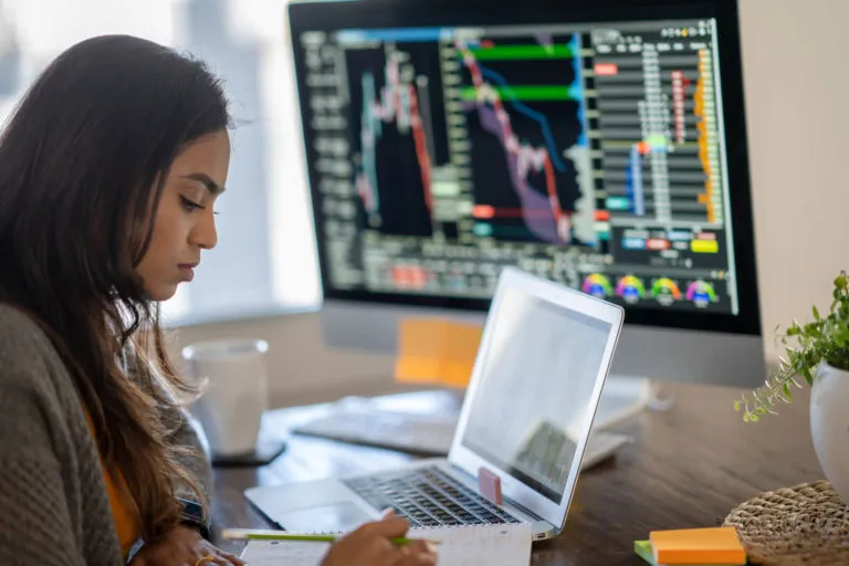 A woman focused on her work at a desk with an open laptop and a large monitor showing financial charts and data behind her.