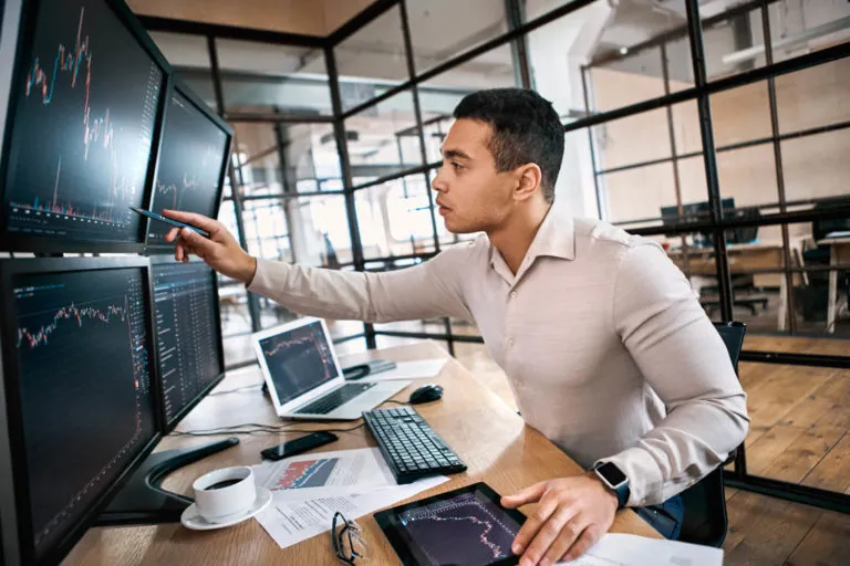 Man sitting at a desk in an office with multiple monitors showing stock market charts, pointing at one screen while holding a tablet displaying a similar chart, surrounded by a laptop, keyboard, documents, and a cup of coffee.