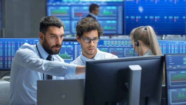 Two men in business attire looking intently at a computer monitor while one of them points at the screen; a woman with a headset is also visible in the background, with multiple monitors displaying financial data and charts.