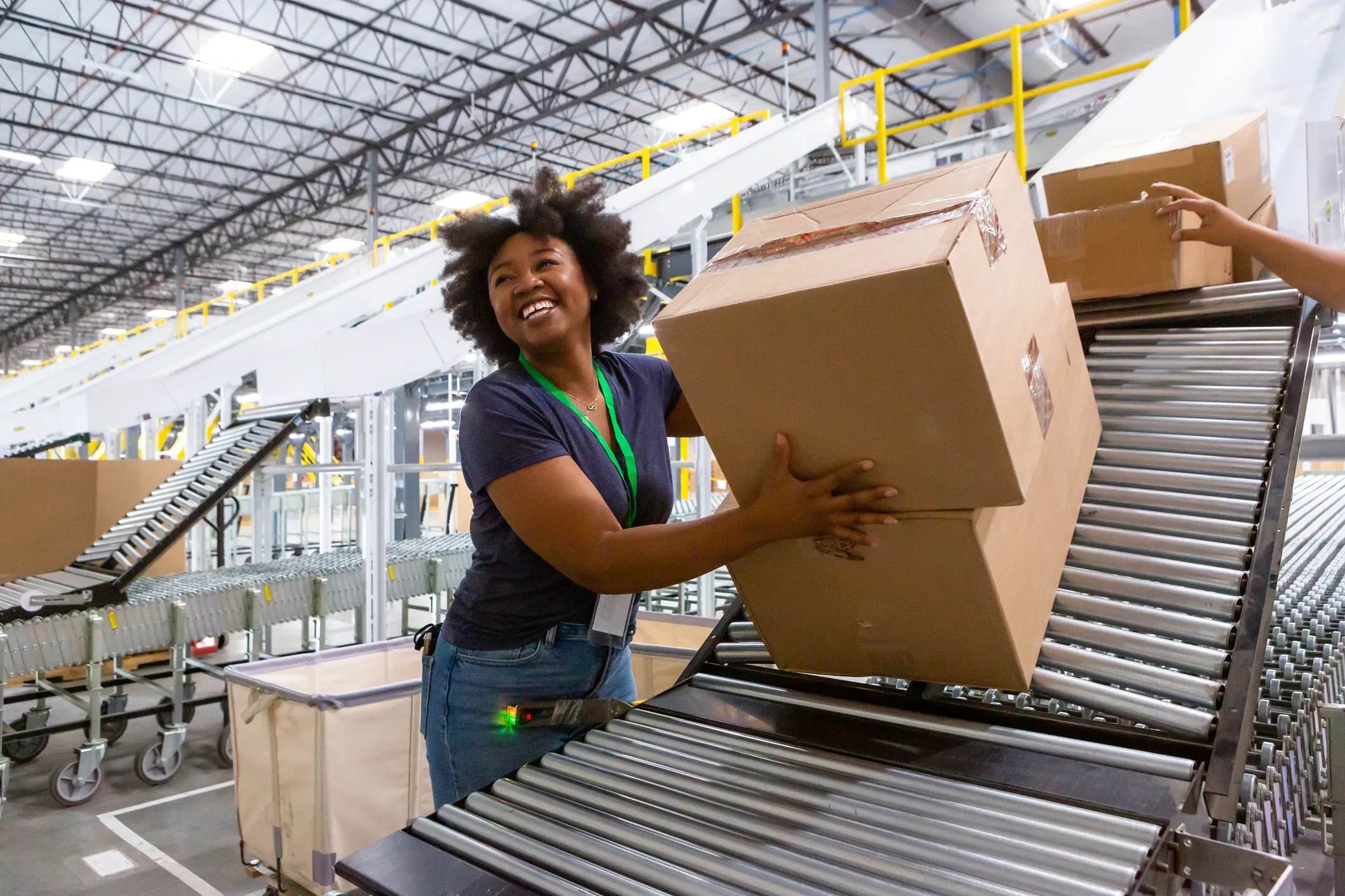 Smiling warehouse worker with curly hair placing a large cardboard box on a metal roller conveyor inside a spacious, well-lit warehouse.