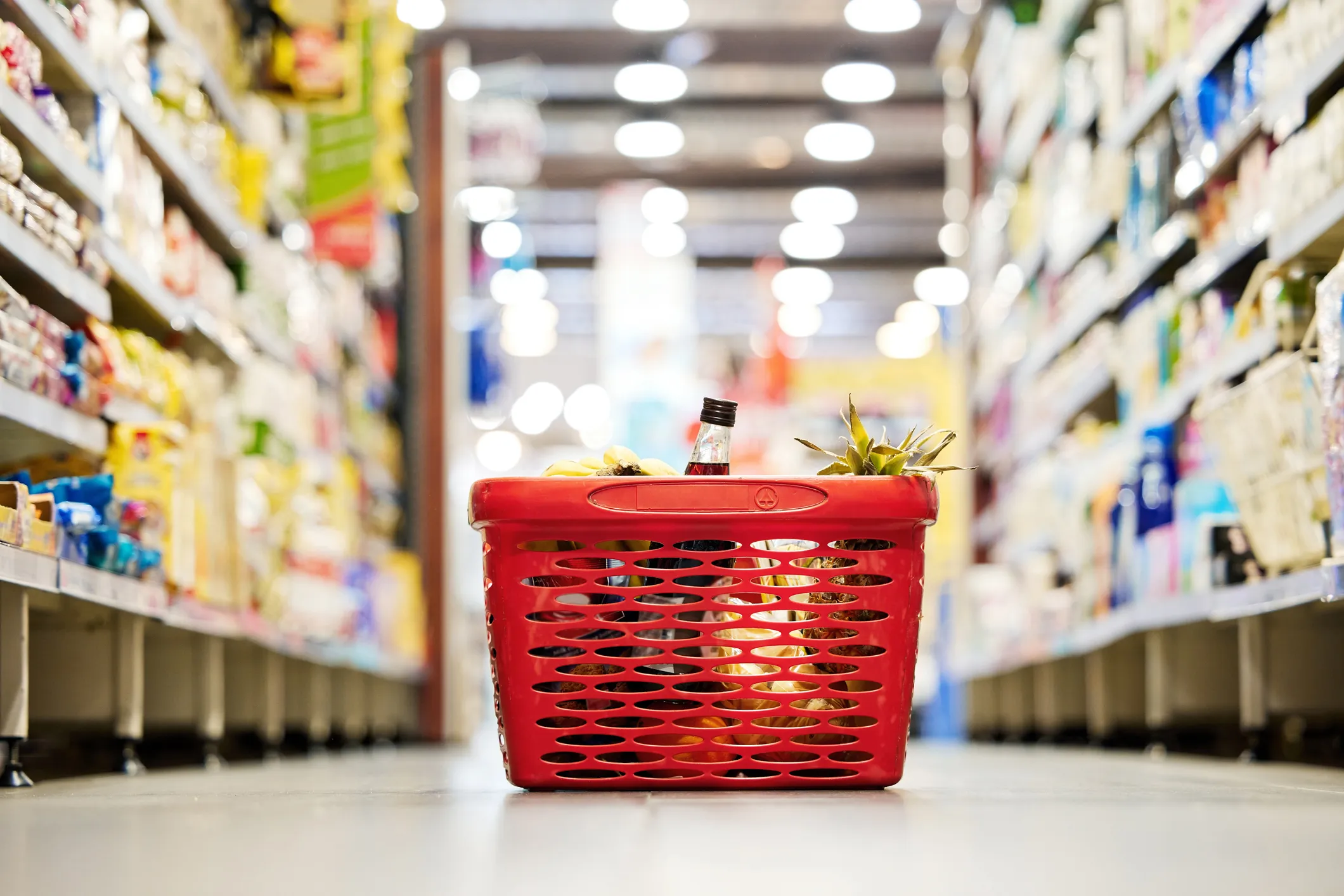Red plastic shopping basket filled with groceries including a pineapple, a bottle, and bananas on the floor aisle in a brightly lit supermarket.