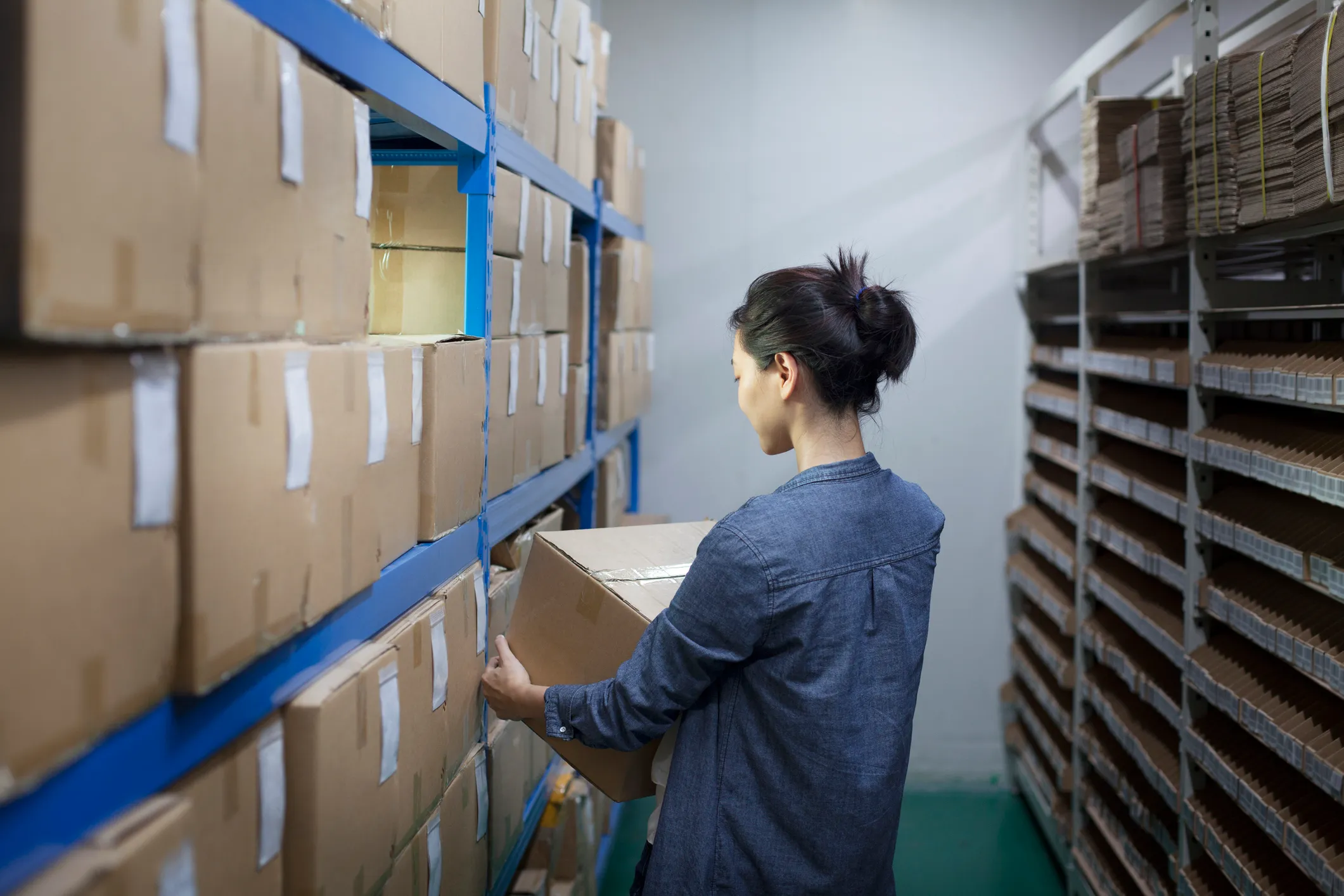 Woman in a blue jacket holding a large cardboard box in a storage room with shelves filled with boxes on both sides.