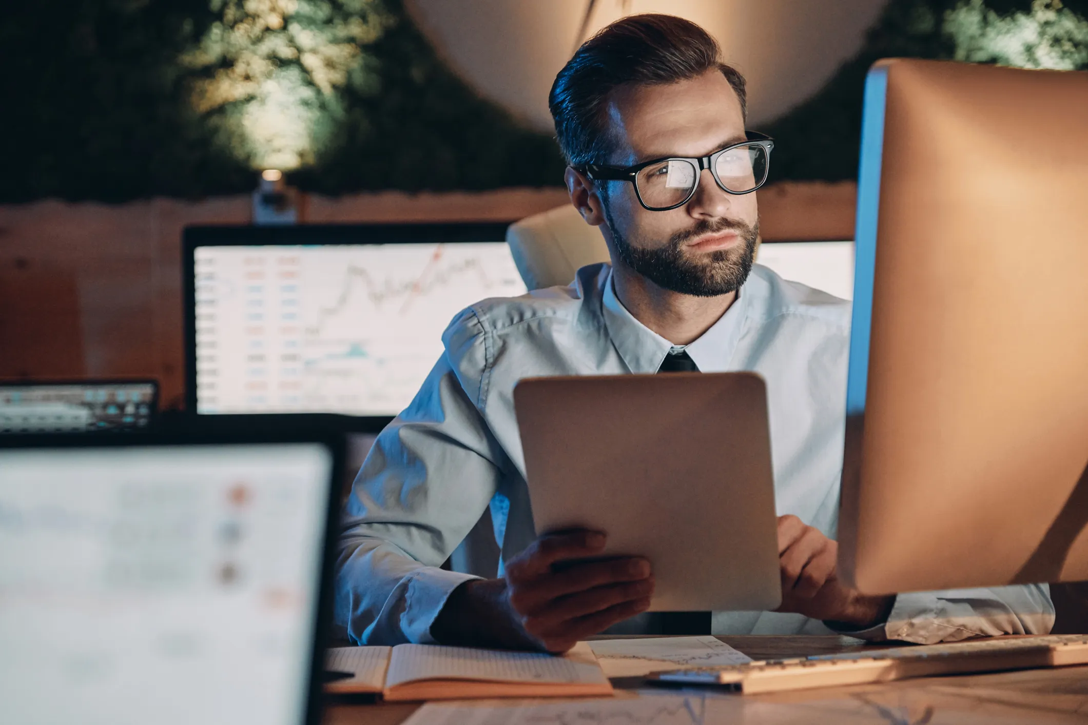 Man with glasses and beard sitting at a desk in a dimly lit office, holding a tablet and looking at a computer monitor displaying financial graphs and data.