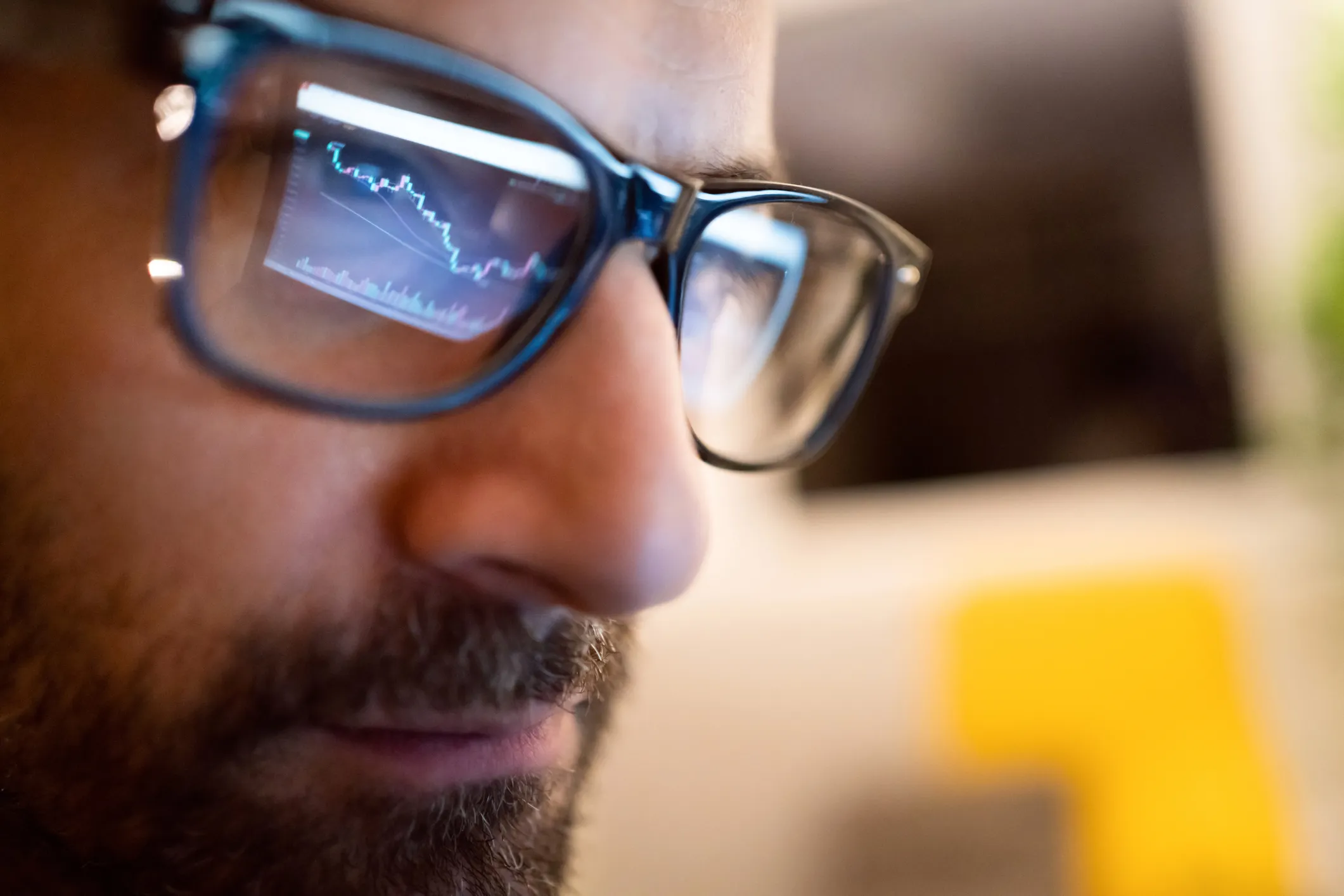 Close-up of a bearded man wearing glasses reflecting a screen showing a declining stock market chart with blue and red lines and a volume bar graph.