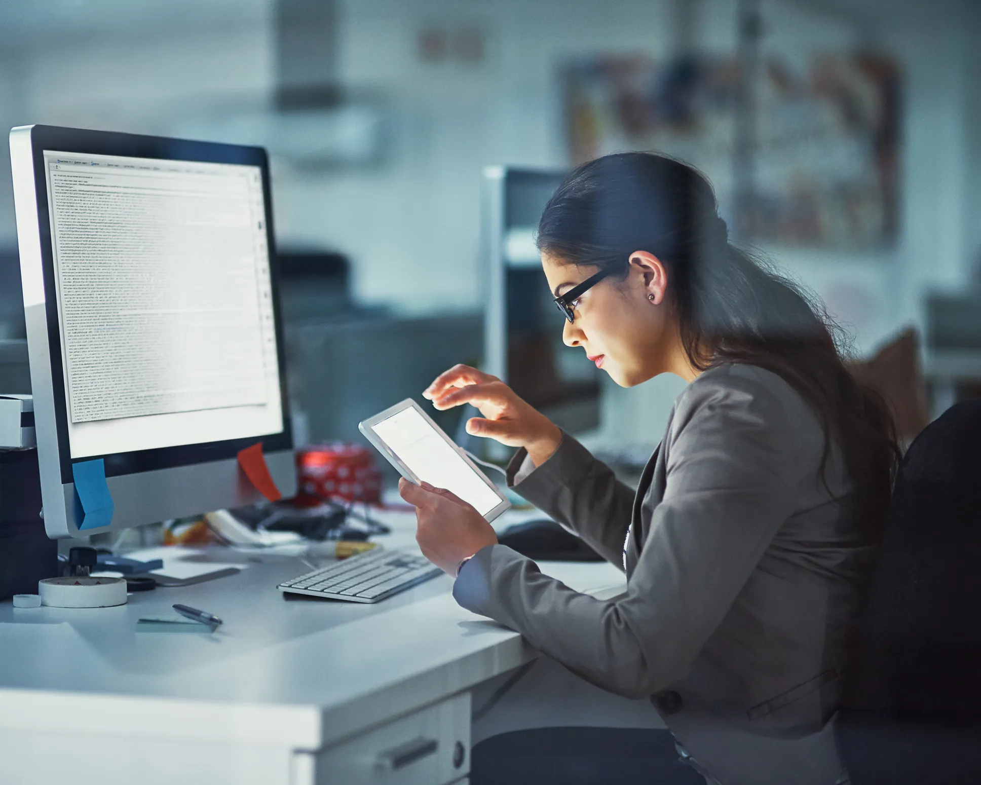 Woman wearing glasses and a blazer working at a white desk using a tablet with a large computer monitor and keyboard nearby in an office setting.