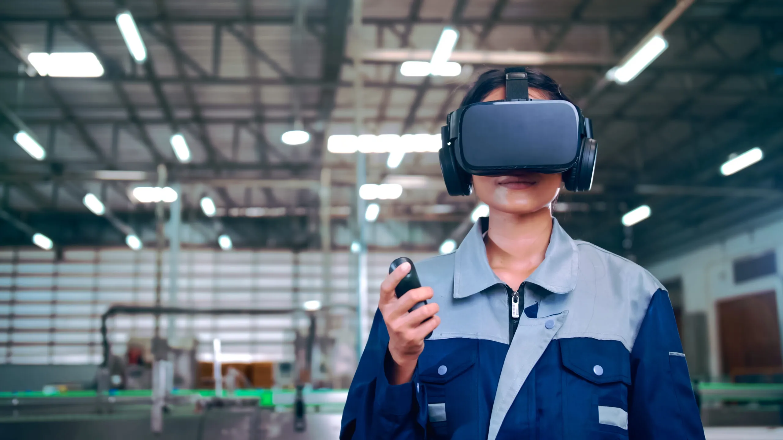 Woman wearing a blue and gray industrial uniform using a virtual reality headset and holding a controller inside a factory or warehouse with blurred machinery in the background.