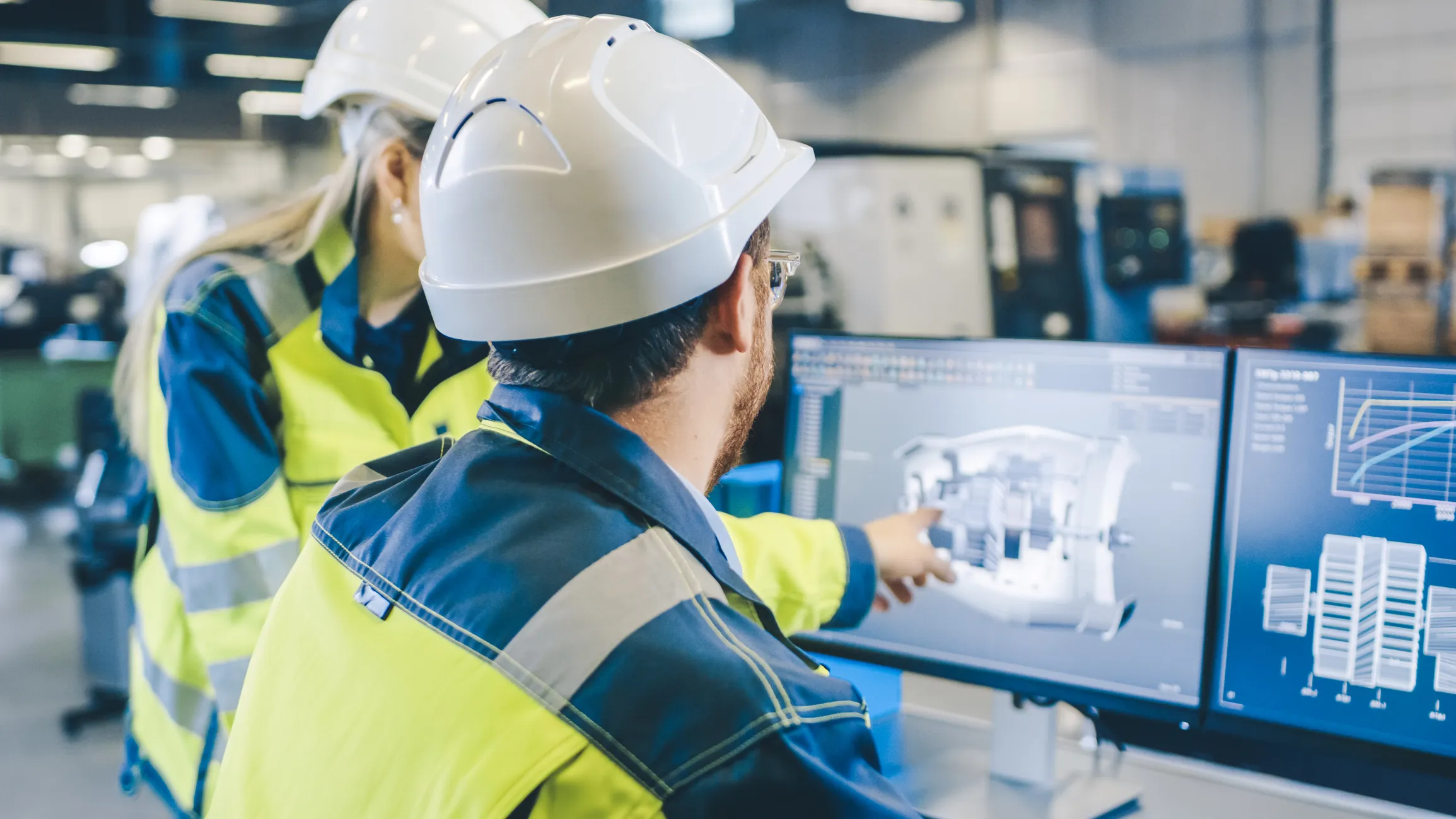 Two engineers wearing white hard hats and yellow safety jackets looking at dual computer monitors displaying a 3D mechanical design and performance graphs inside an industrial facility.