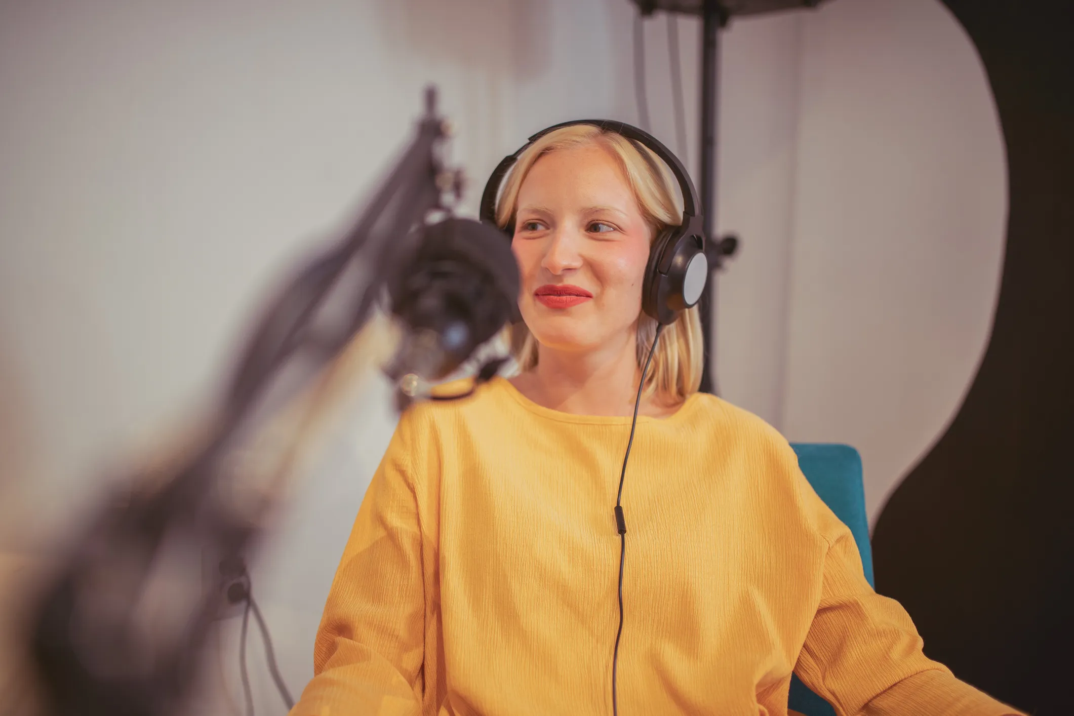 Young woman with blonde hair wearing a yellow top and black headphones, speaking into a microphone in a recording studio setting.