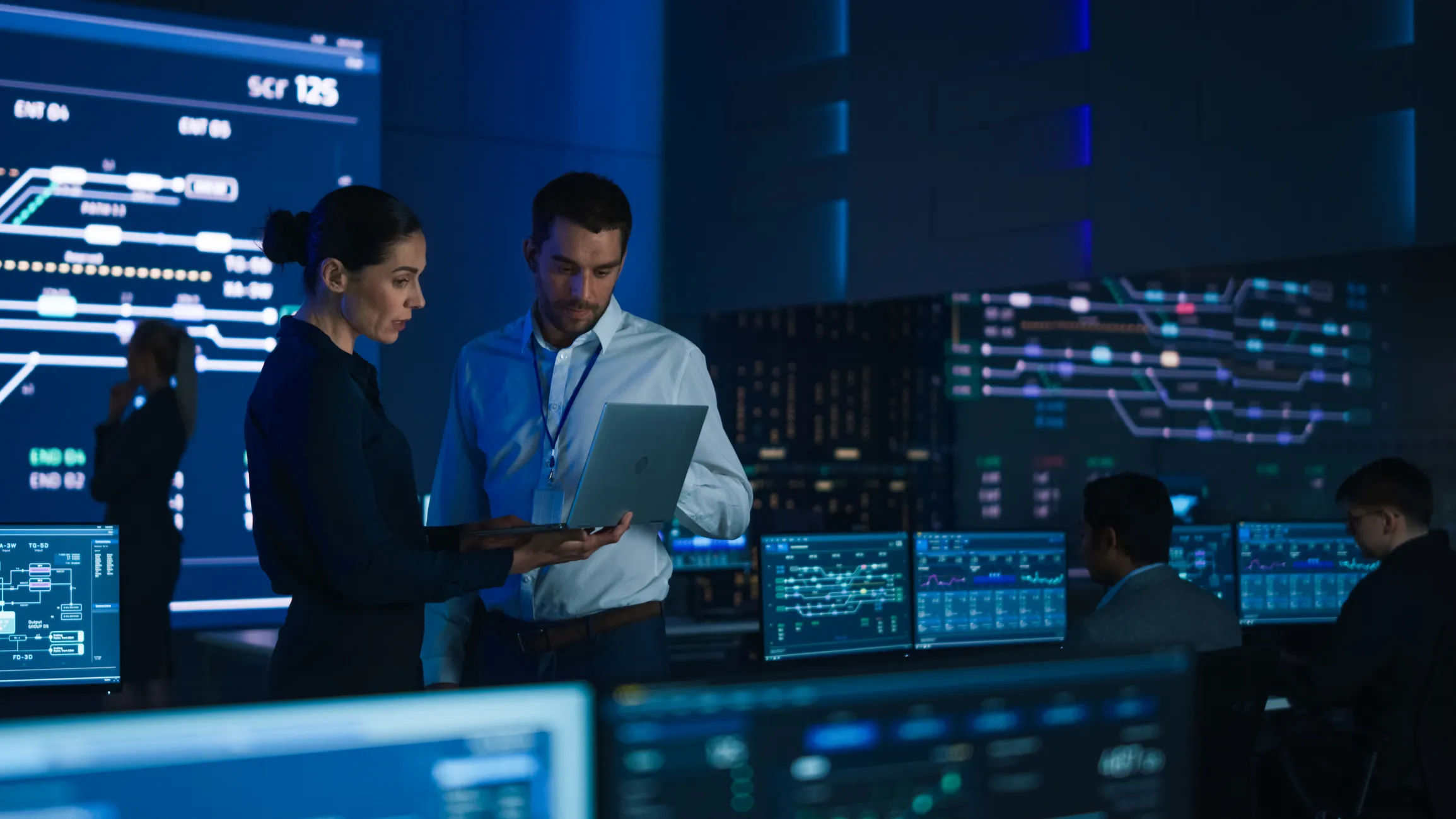 Two professionals standing and discussing information on a laptop inside a dark control room with multiple large monitors displaying technical graphs and data, while two other people work at desks with multiple screens in the background.