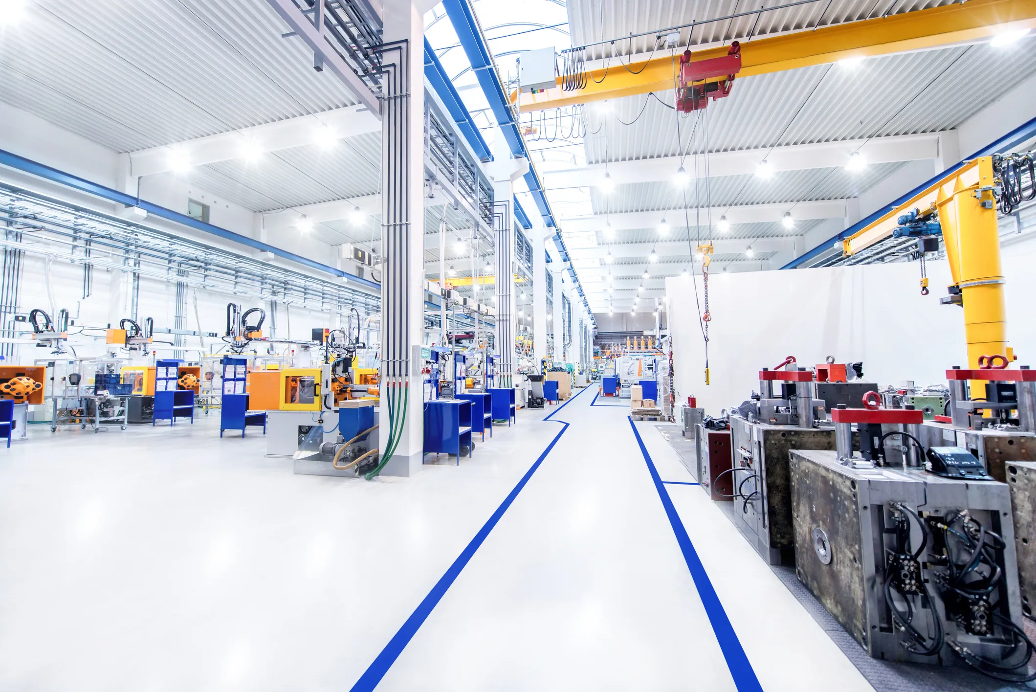 Bright, spacious industrial factory interior with high ceiling, rows of machinery on both sides, blue safety lines on floor, and overhead cranes.