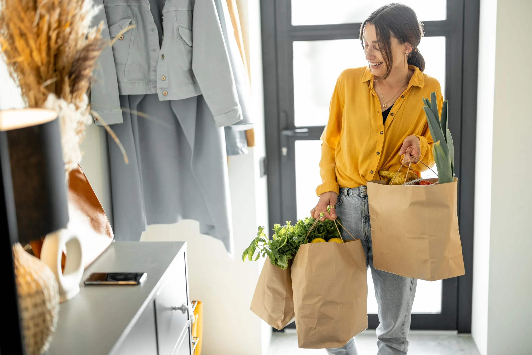 Woman wearing a yellow shirt and jeans carrying two brown paper grocery bags filled with fresh vegetables indoors near a door, smiling and looking down.