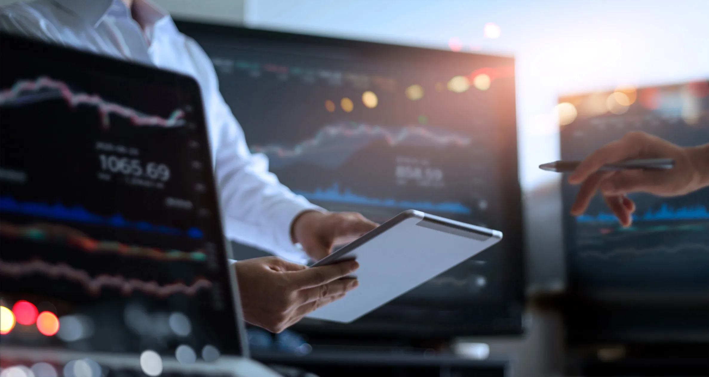 Person holding a tablet with financial charts displayed on multiple monitors in the background, showing stock market data and graphs.