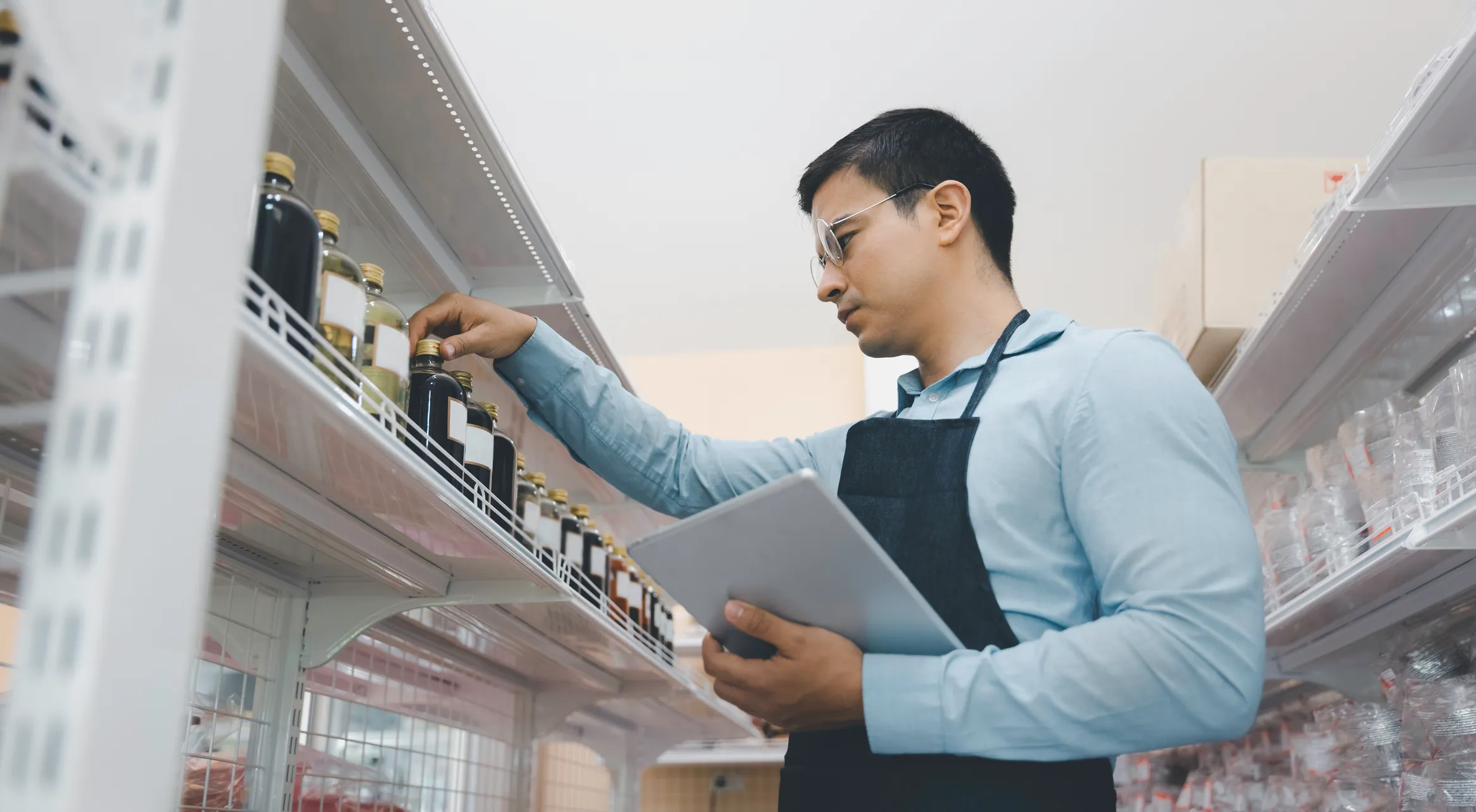 Man wearing glasses and a black apron checking bottles on a store shelf while holding a tablet.