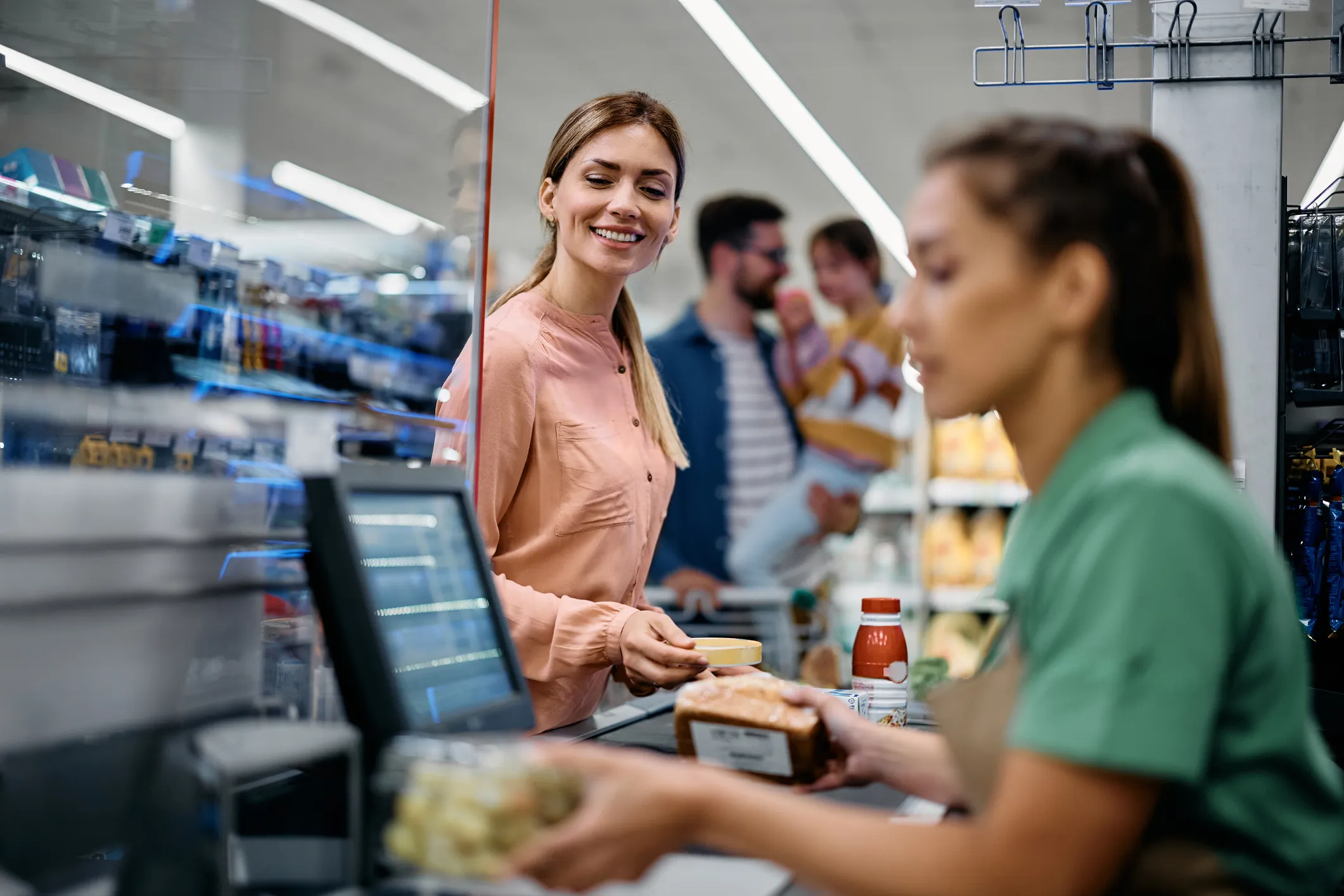 Smiling woman in a pink blouse paying for groceries at a store checkout counter with a cashier in green uniform scanning items and a man holding a child in the background.