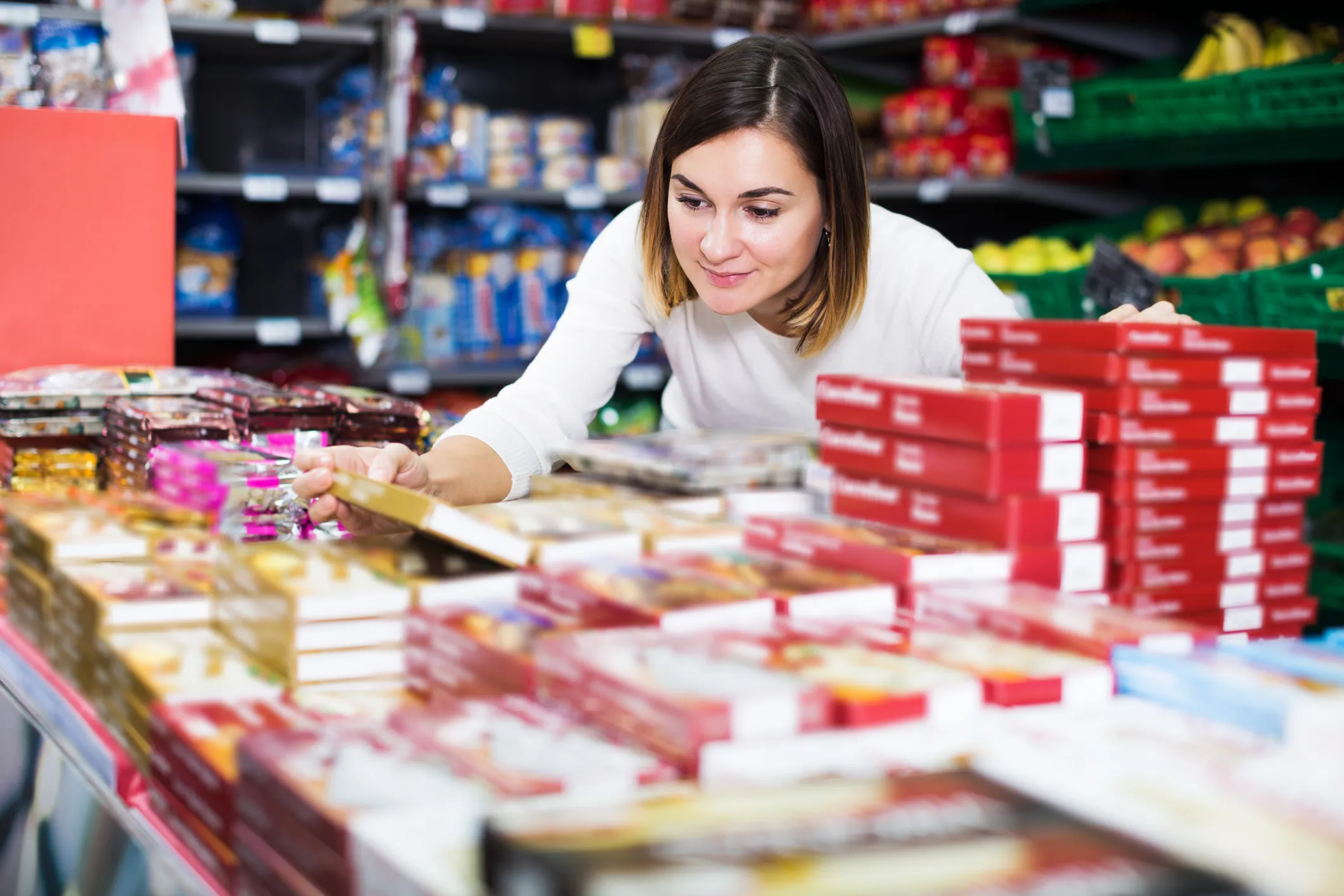 A woman in a white sweater selecting a packaged food item from a supermarket aisle filled with stacks of various boxed goods and fresh produce in the background.