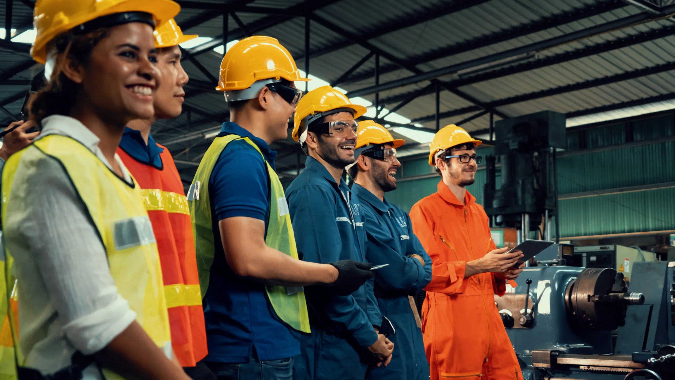 A diverse group of industrial workers and engineers wearing safety helmets, protective glasses, and vests gathered in a manufacturing facility, smiling and engaged, with machinery in the background.